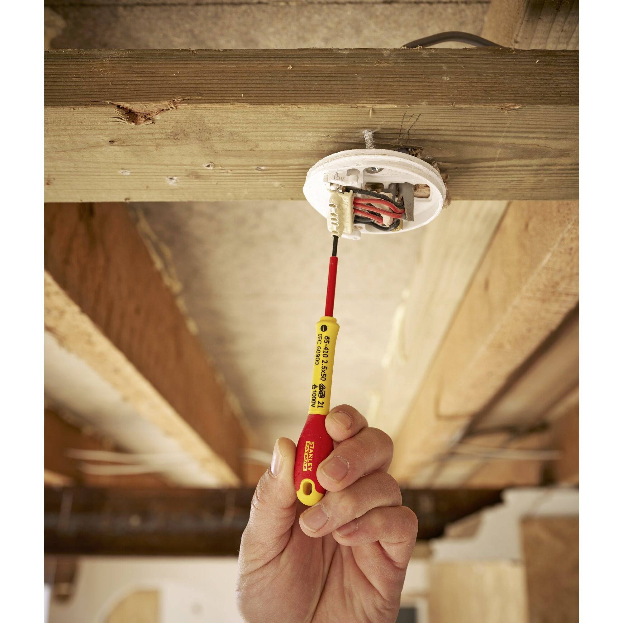 Une main tient un testeur de tension sur un branchement électrique dans un plafond en bois. L'accent est mis sur la sécurité lors des installations électriques.