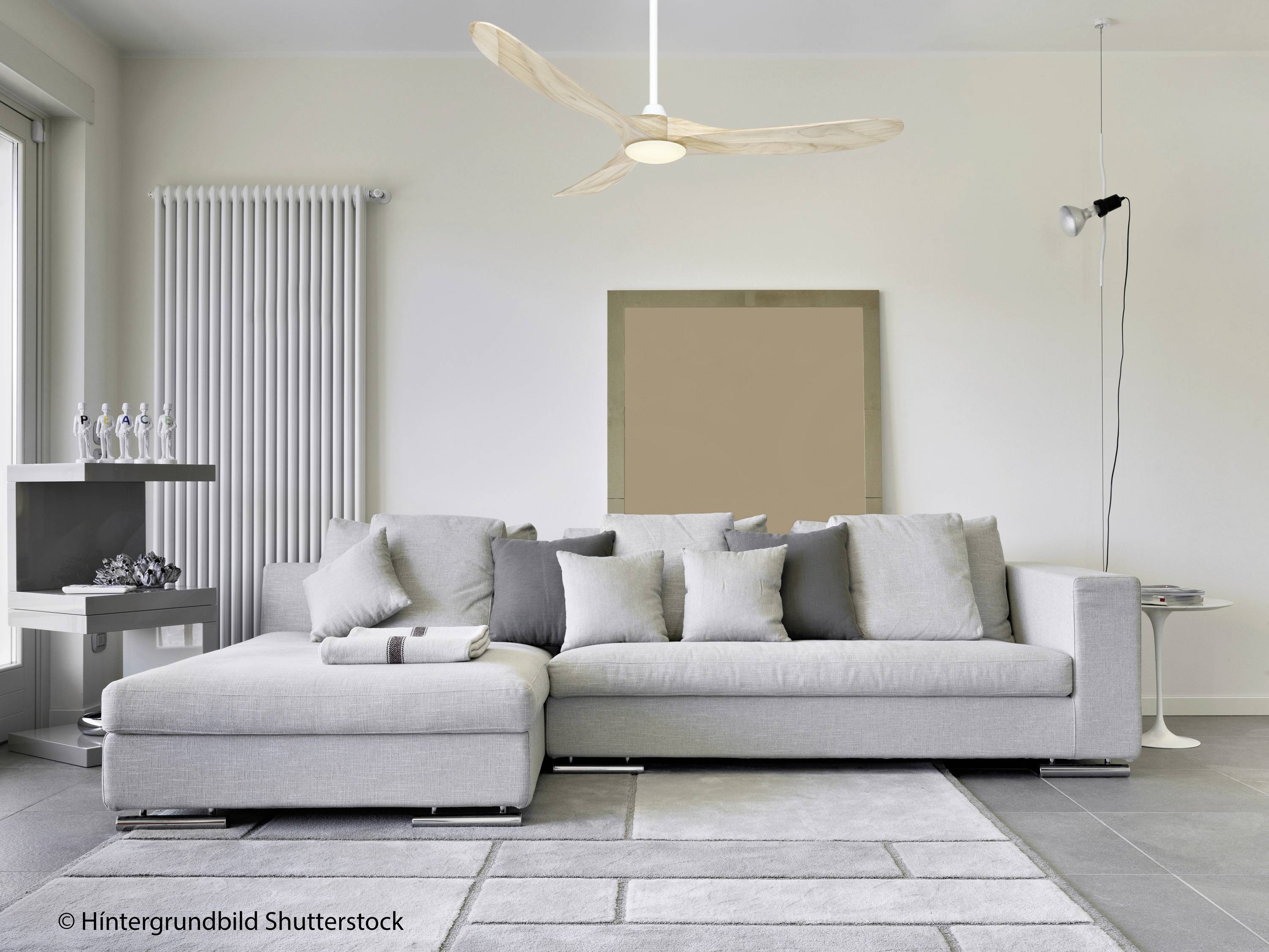 Salon moderne avec canapé d'angle gris, coussins décoratifs, tapis, parquet clair, ventilateur de plafond et éclairage minimaliste.