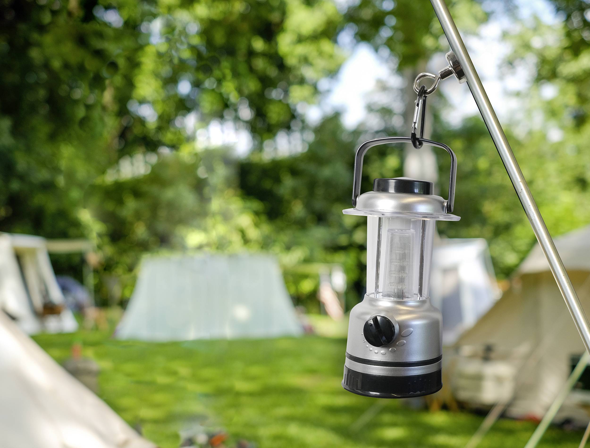 Une lanterne argentée est suspendue à un mât de tente dans un espace vert. En arrière-plan, plusieurs tentes sont visibles de manière floue.