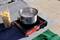 Une casserole avec de l'eau bouillante et des pâtes est posée sur un réchaud à gaz portable à l'extérieur. À côté se trouve un paquet de pâtes ouvert.
