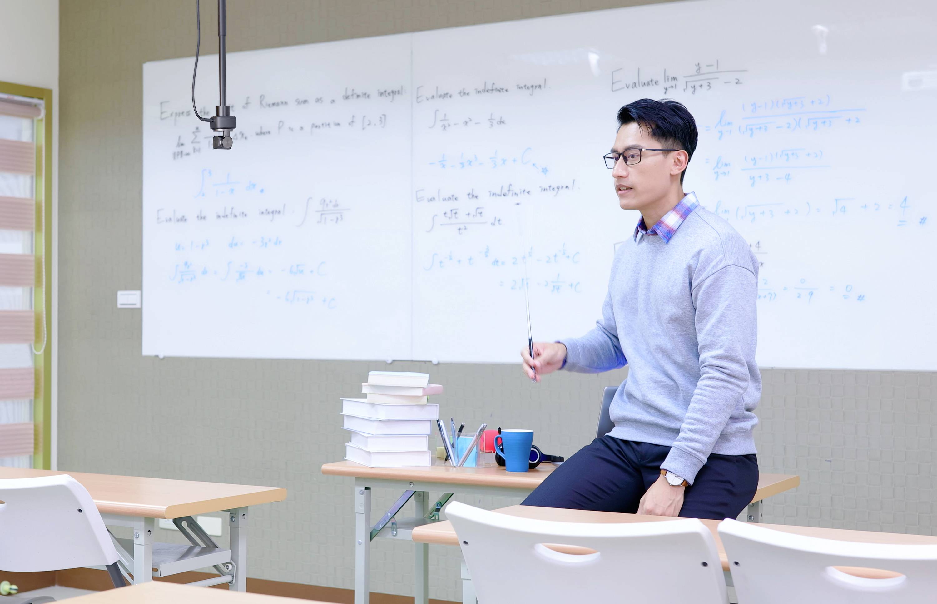 Un homme est assis sur une table dans une salle de classe, entouré de livres empilés. Au fond, un tableau noir est couvert de formules mathématiques.
