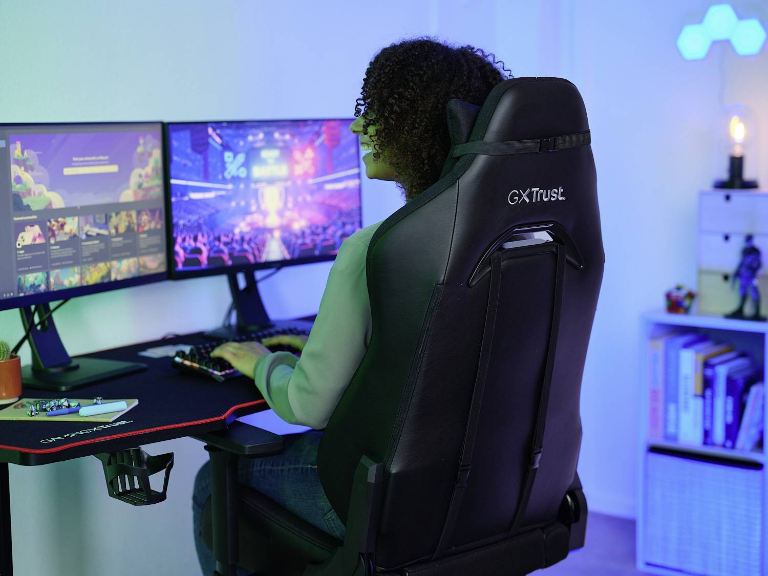 Une personne est assise à un bureau avec deux grands écrans et joue à un jeu vidéo. La pièce est éclairée en bleu.