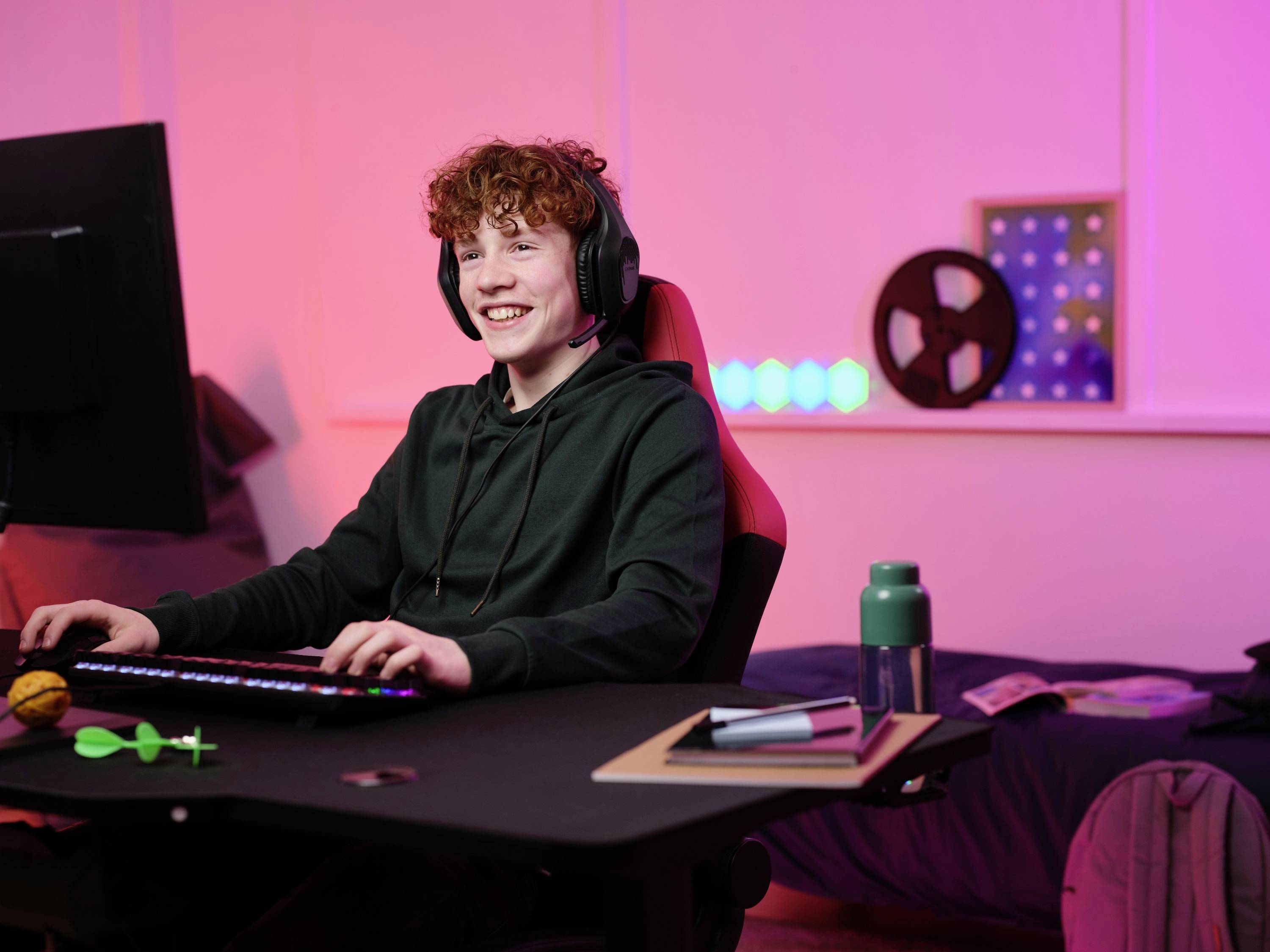 Un adolescent est assis à un bureau, portant des écouteurs et souriant tout en jouant sur son ordinateur. Un étagère éclairée est visible en arrière-plan.
