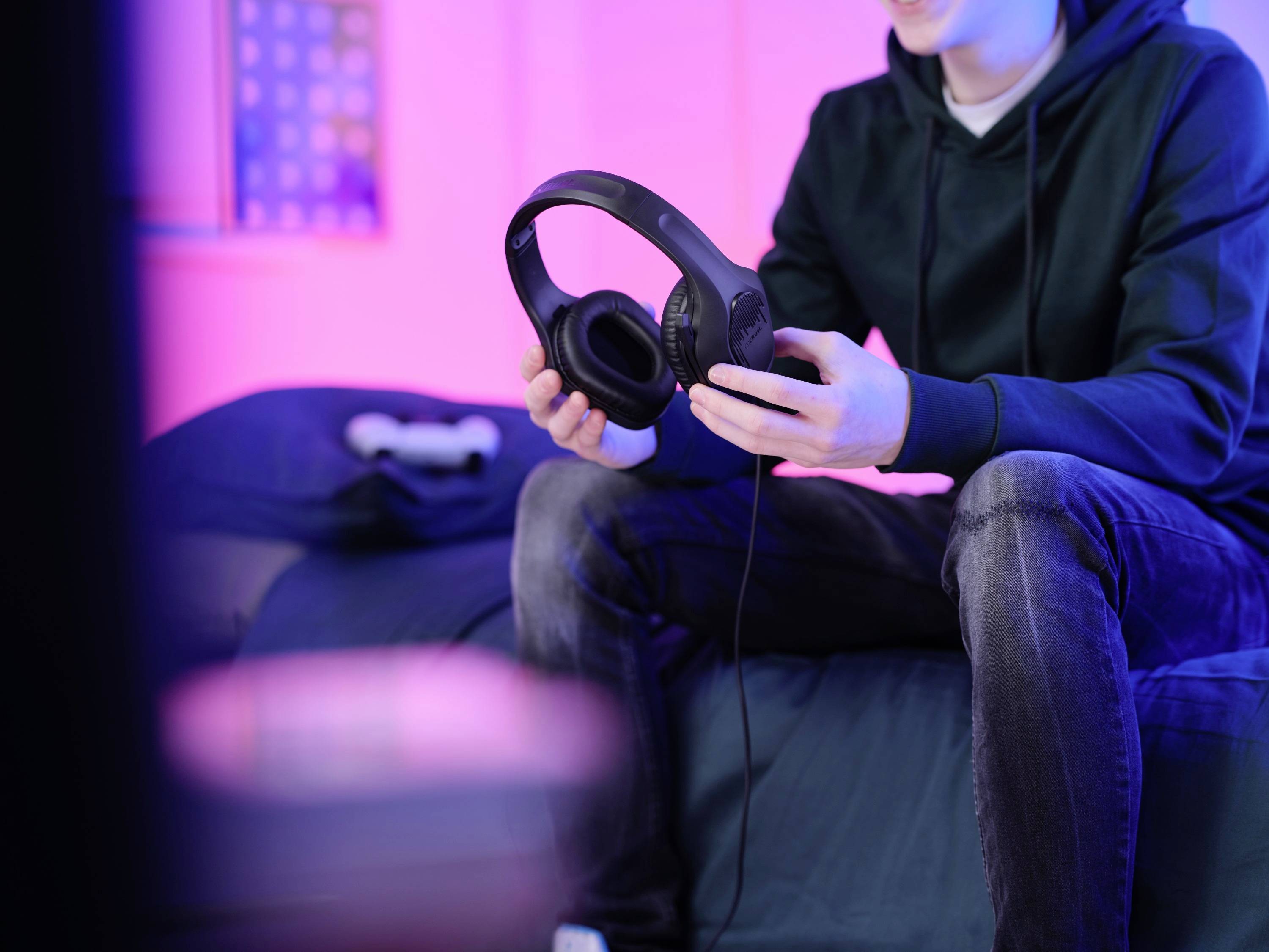 Une personne est assise sur un lit dans une pièce éclairée en rose, tenant un casque noir. Un contrôleur est visible en arrière-plan.