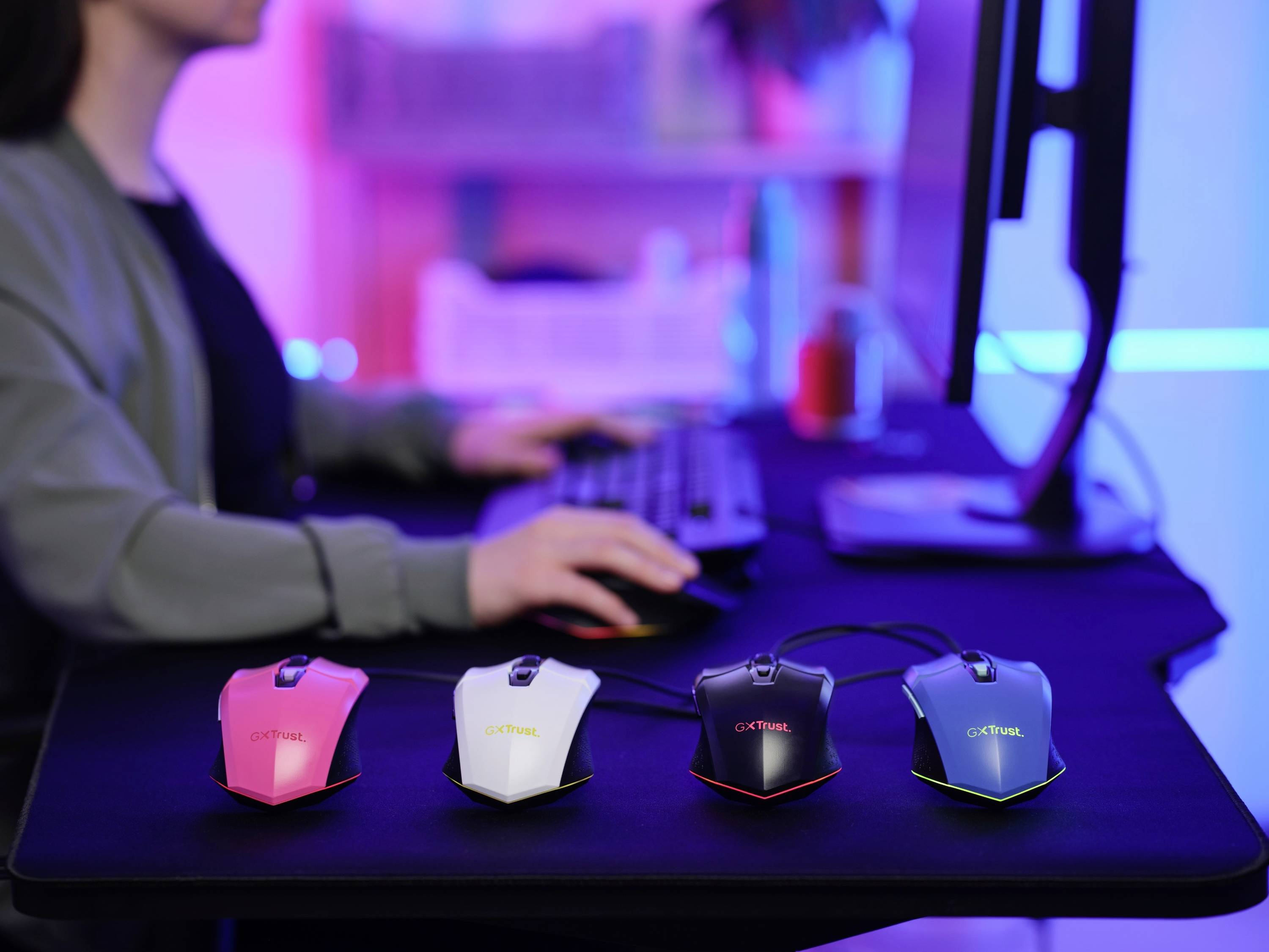 Une personne travaille sur un ordinateur. Devant elle sont posées quatre souris gaming de différentes couleurs : rose, blanche, noire et bleue.