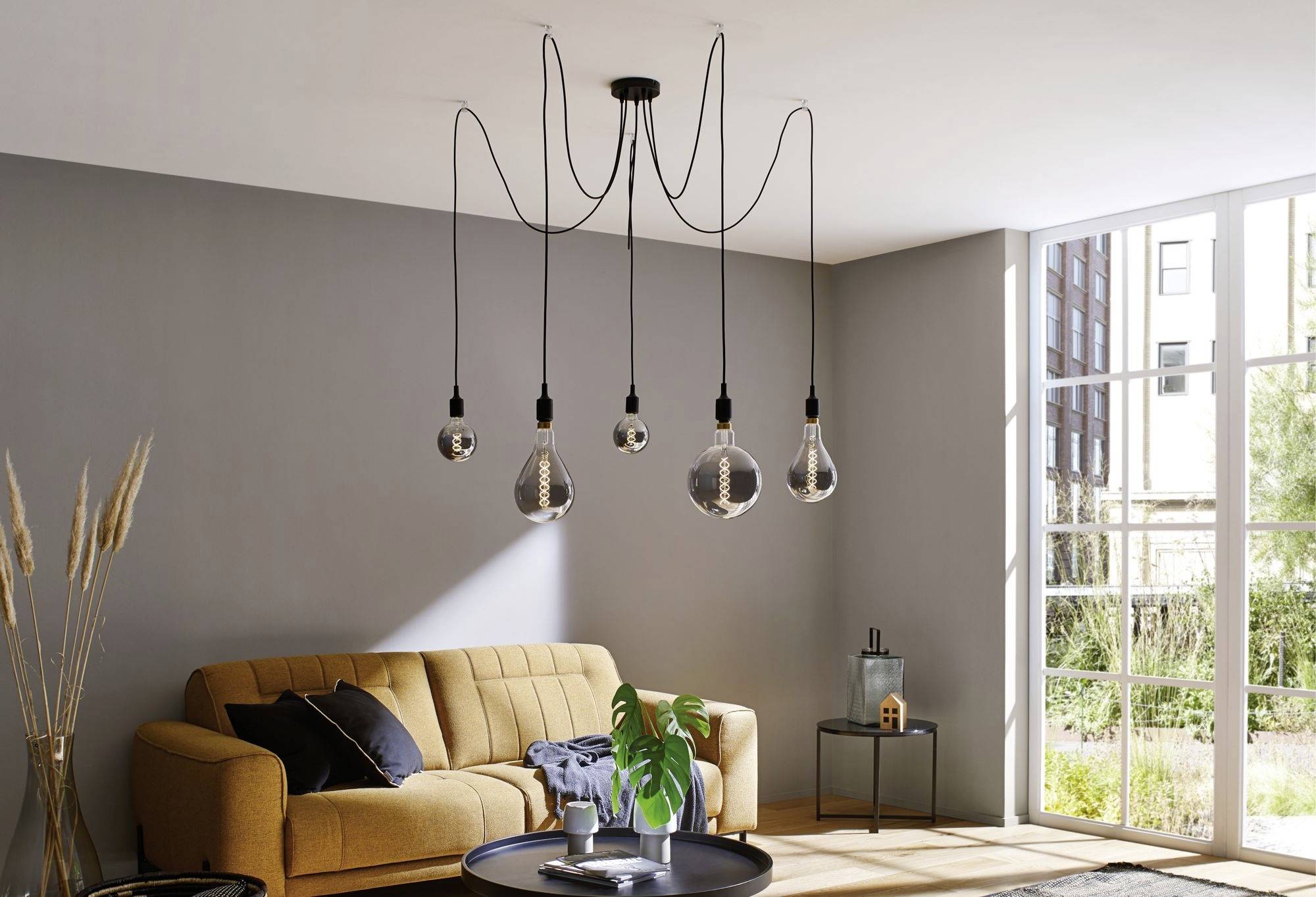 Salon moderne avec canapé jaune, table basse en verre et suspension avec plusieurs ampoules. Une grande fenêtre laisse entrer la lumière du jour.