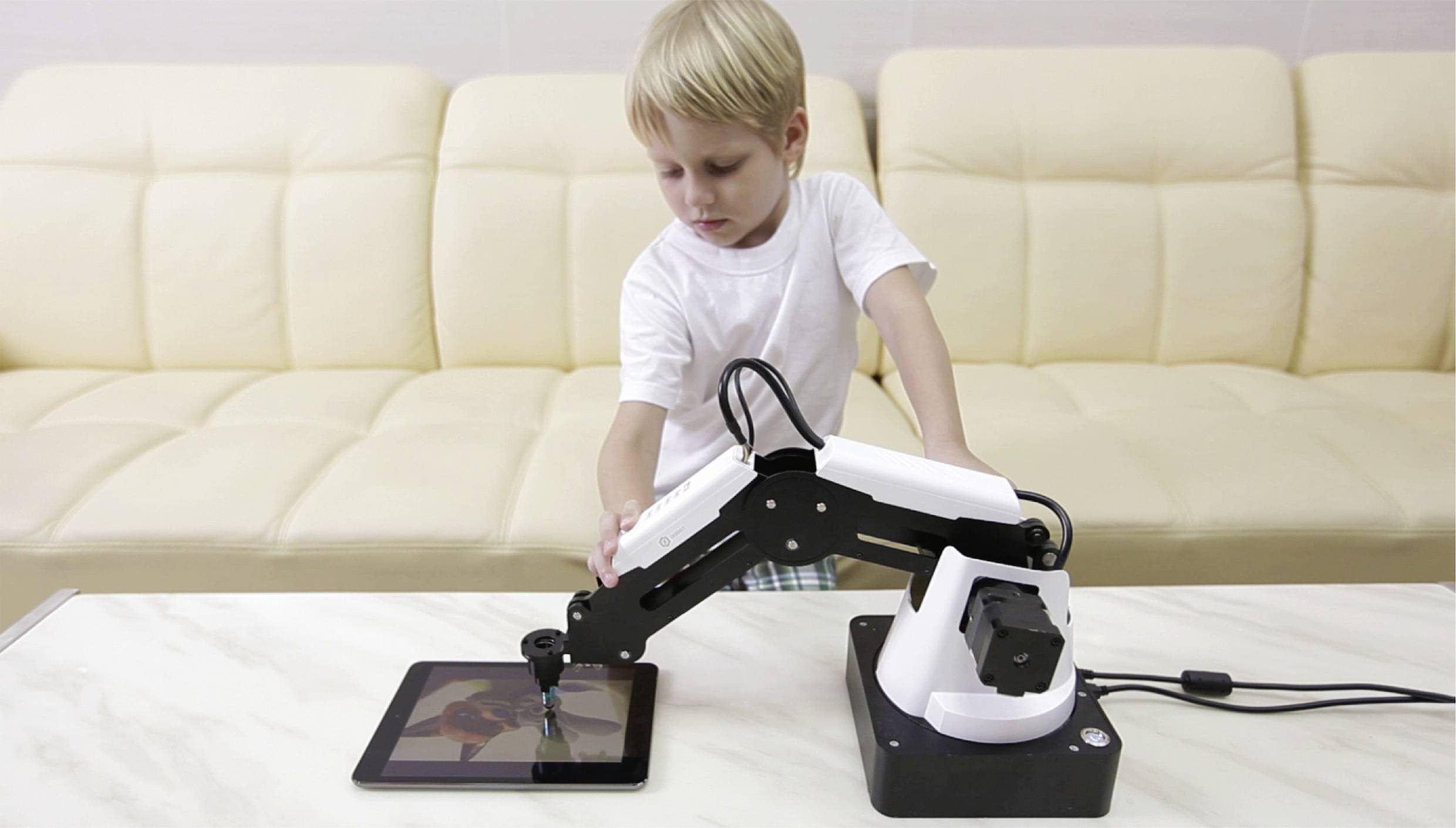 Un enfant commande un bras robotique qui dessine sur une tablette. Un canapé beige est visible en arrière-plan.