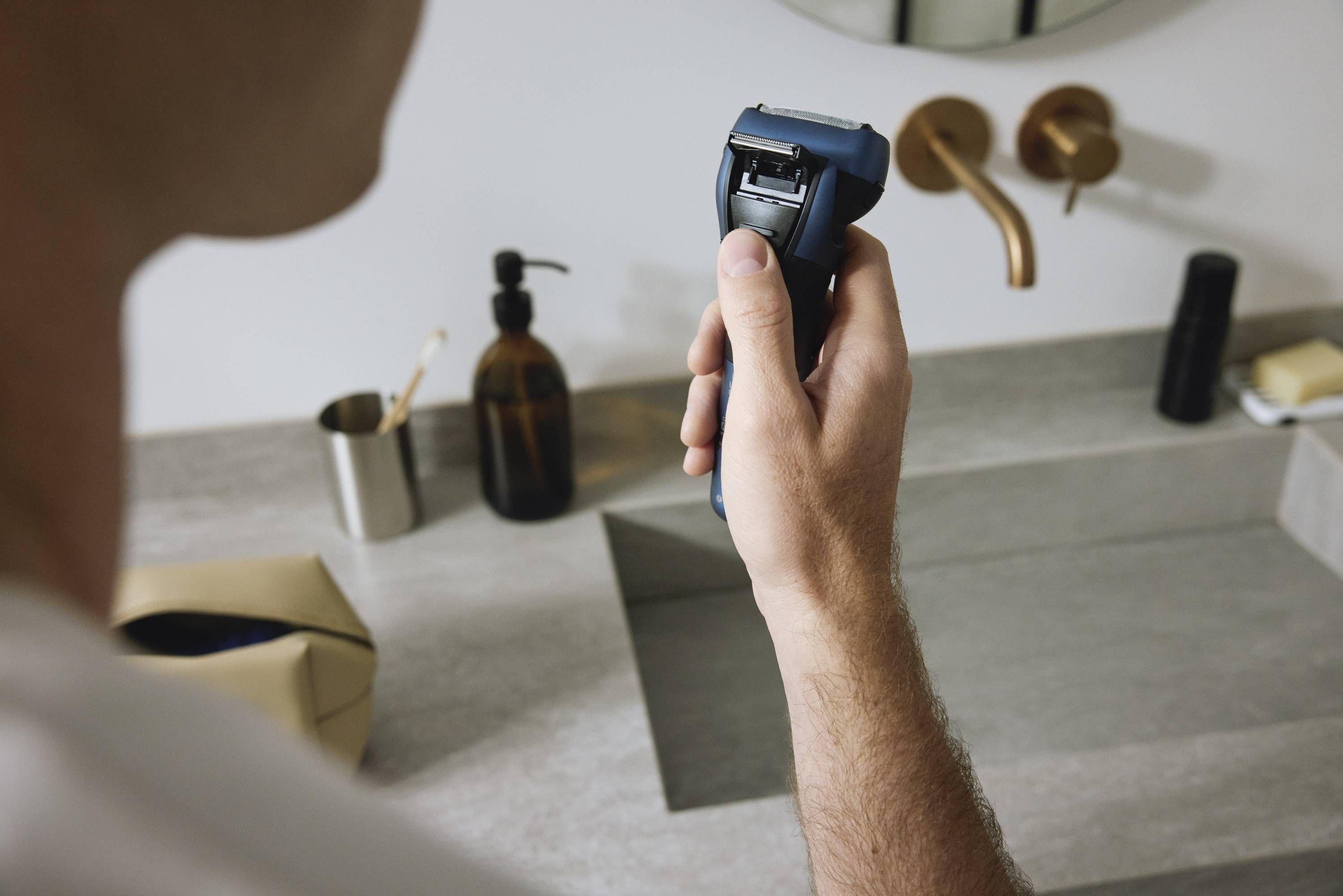 Une personne tient un rasoir électrique au-dessus d'un lavabo dans une salle de bain moderne. Des distributeurs de savon et des robinets sont visibles en arrière-plan.