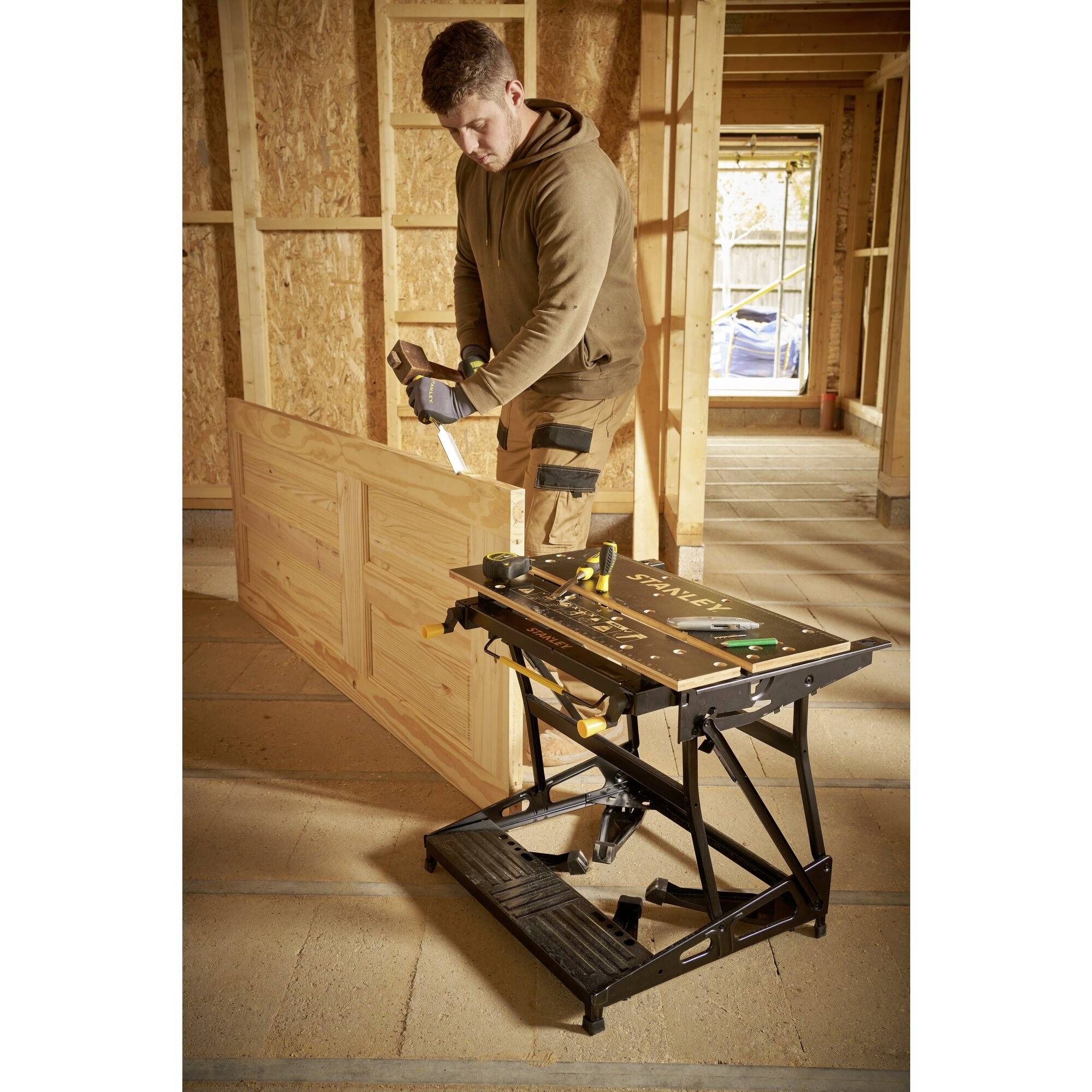 Un homme en vêtements de travail scie une planche de bois avec une scie à main dans un atelier. Un établi avec des outils est situé à droite.