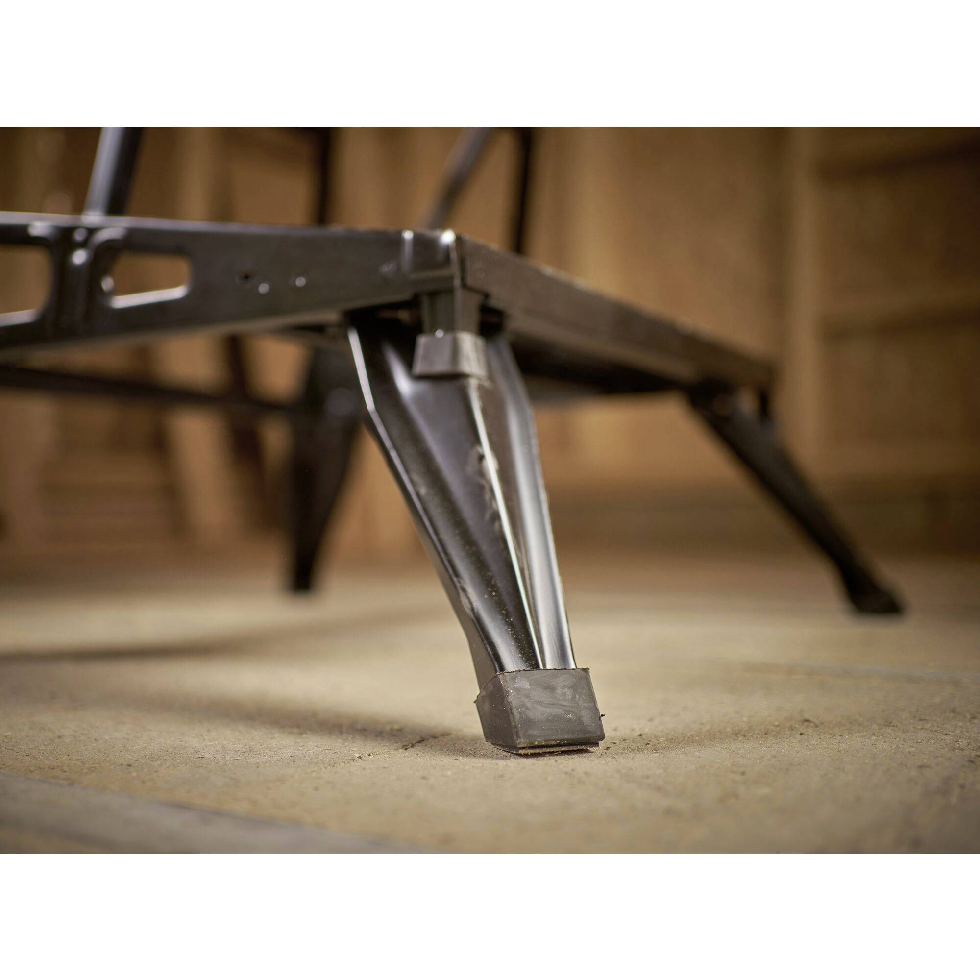 Une jambe de table en métal sur un sol poussiéreux, partiellement cachée par un bord de table, avec un espace en bois flou en arrière-plan.