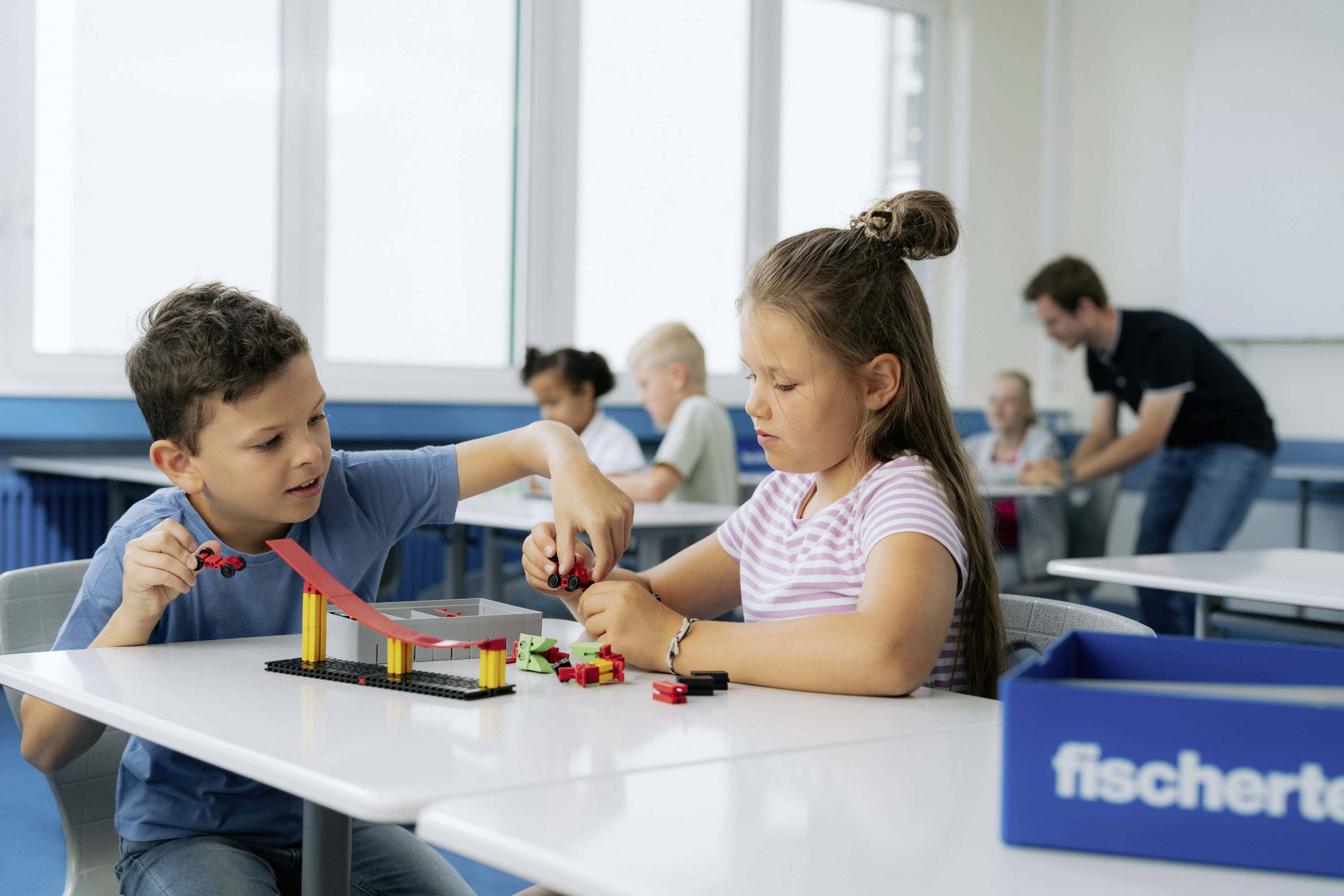 Des enfants construisent avec des jouets de construction autour d'une table. D'autres enfants travaillent en arrière-plan. Une boîte de pièces est visible.