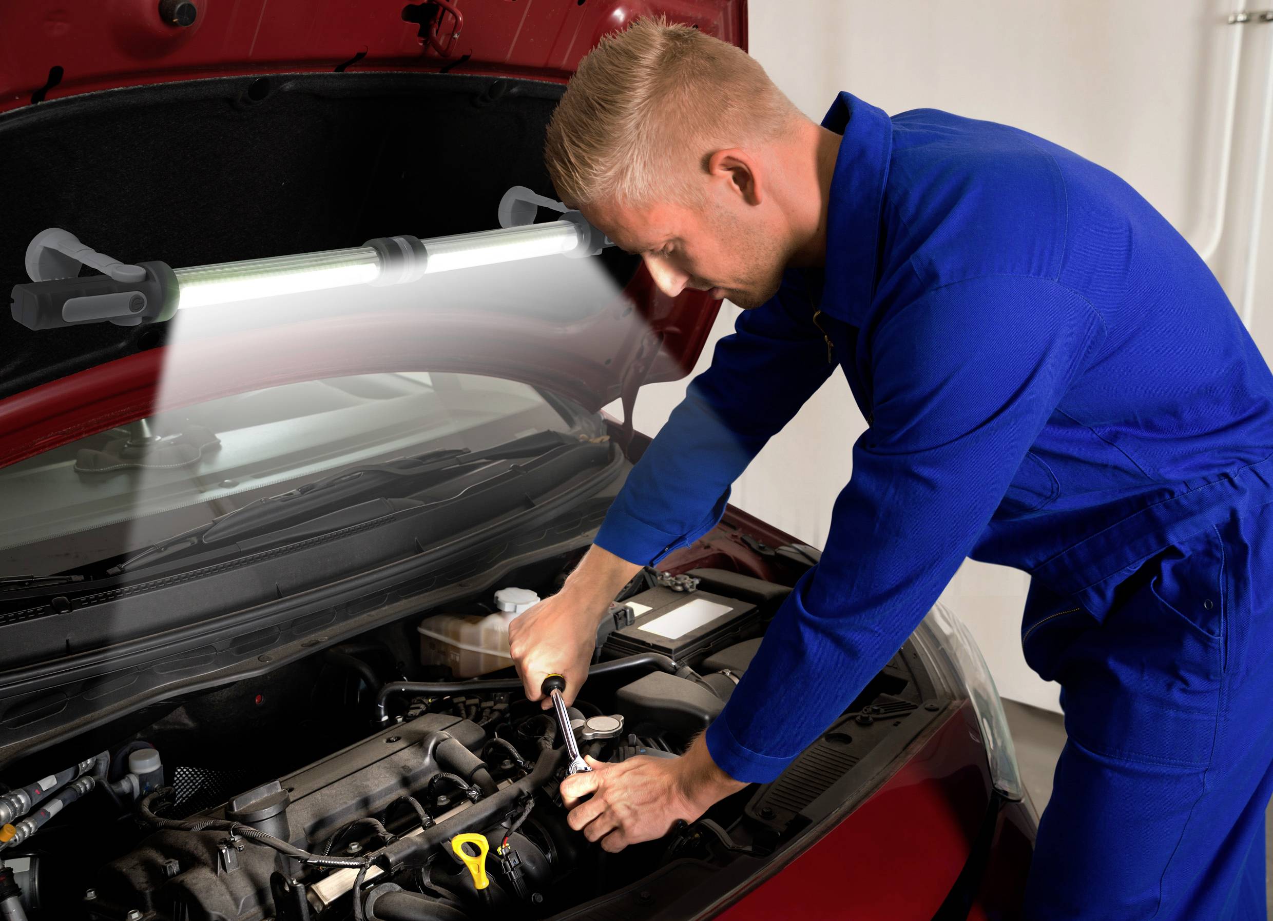 Un mécanicien en bleu de travail répare le moteur d'une voiture dans un atelier. Une lampe éclaire le compartiment moteur.