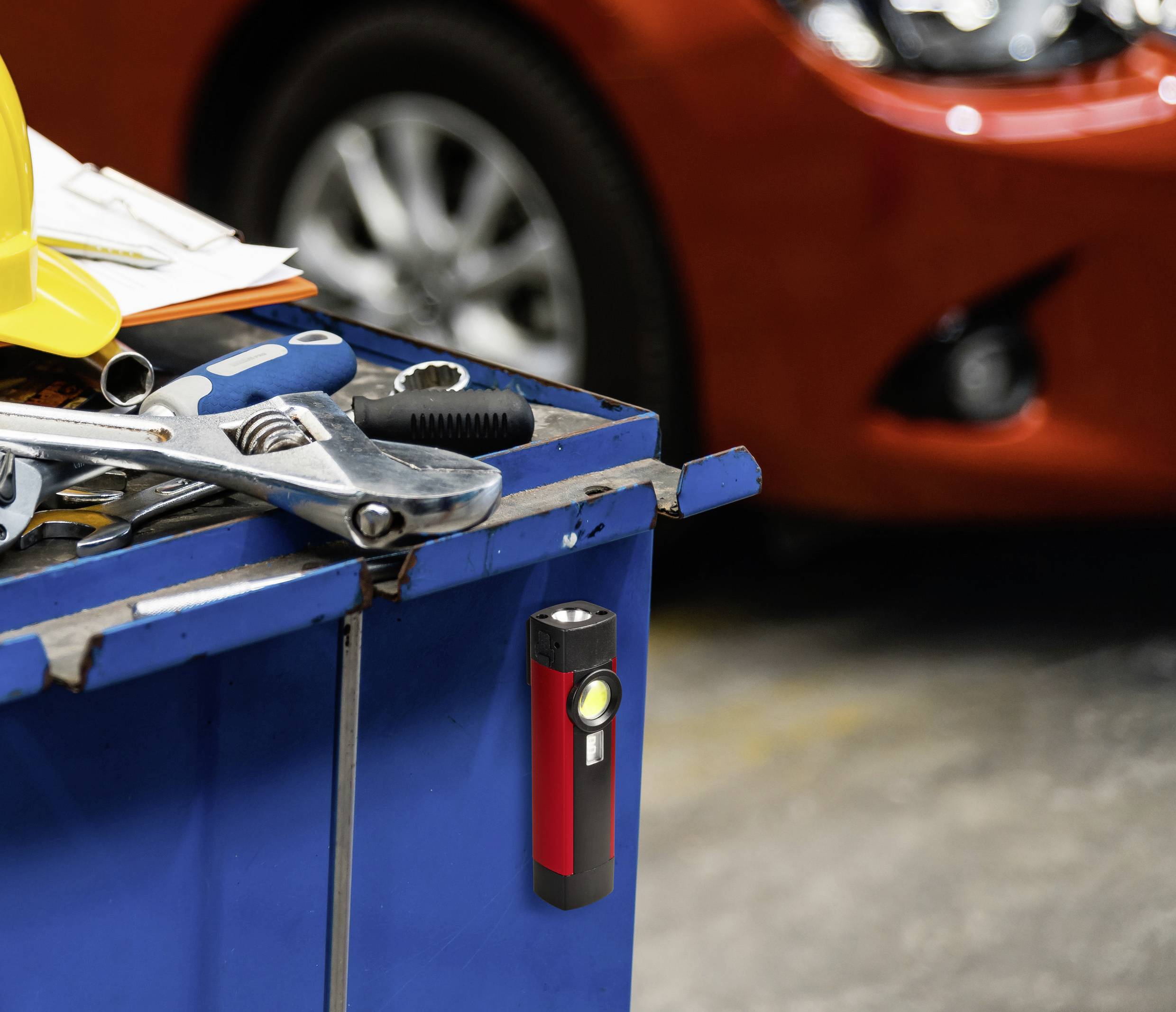 Une lampe de poche est suspendue à un chariot à outils dans un atelier. À l'arrière-plan, la roue avant floue d'une voiture rouge est visible.