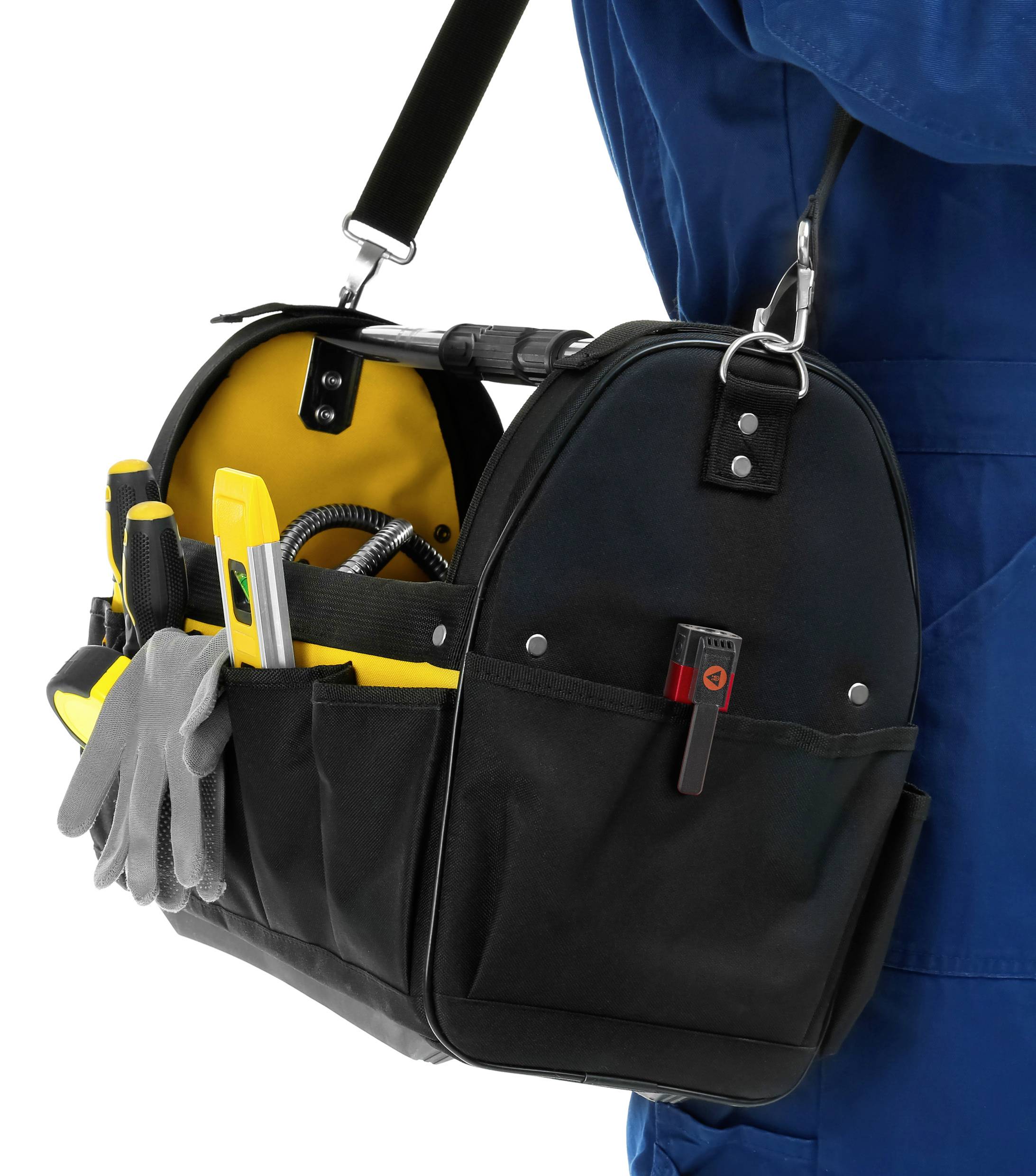 Un artisan en tenue de travail bleue porte un sac à outils noir rempli d'outils manuels, tels que des tournevis et des pinces.