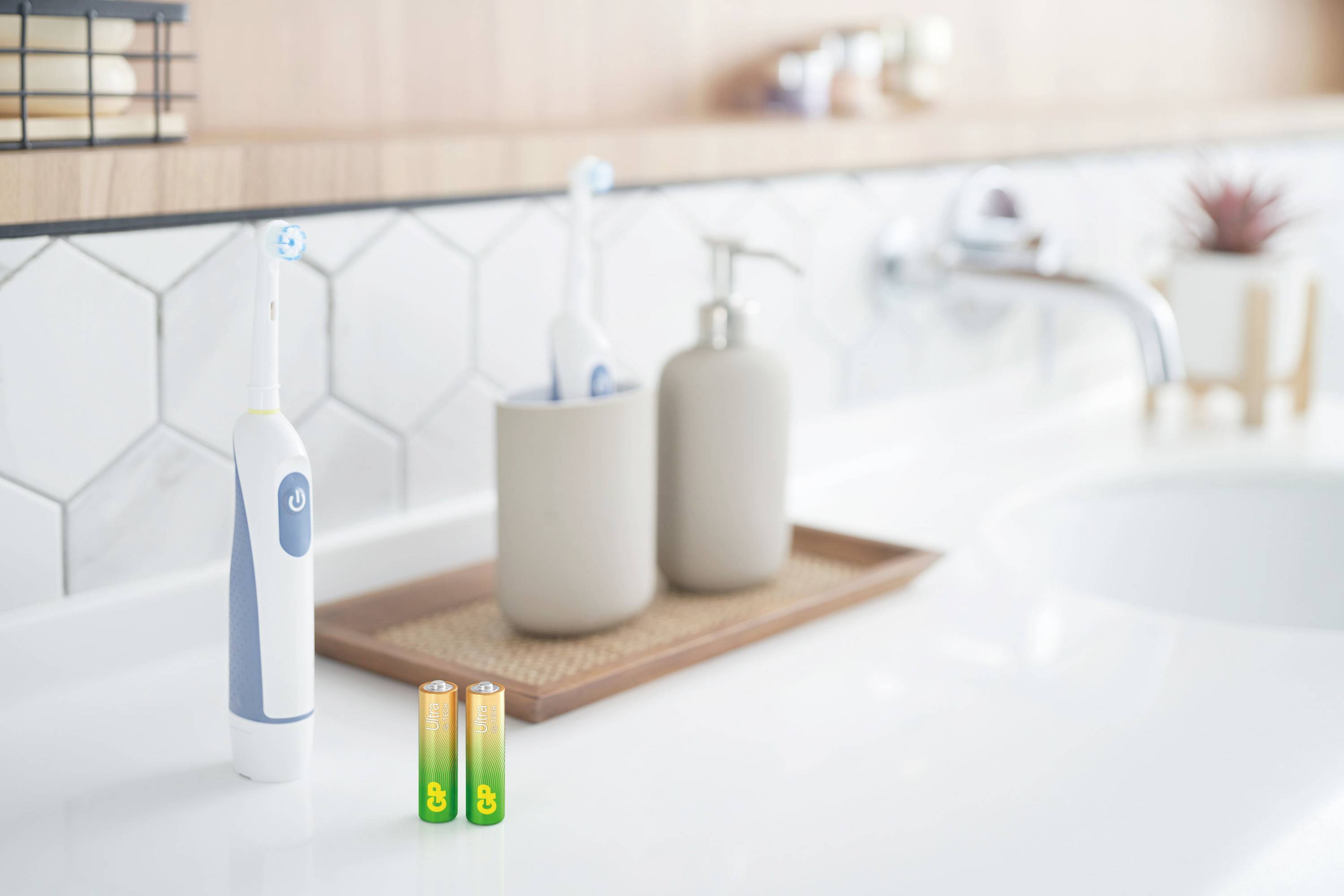 Brosse à dents électrique et piles sur un lavabo blanc. En arrière-plan, un gobelet à brosse à dents avec une deuxième brosse et un distributeur de savon.
