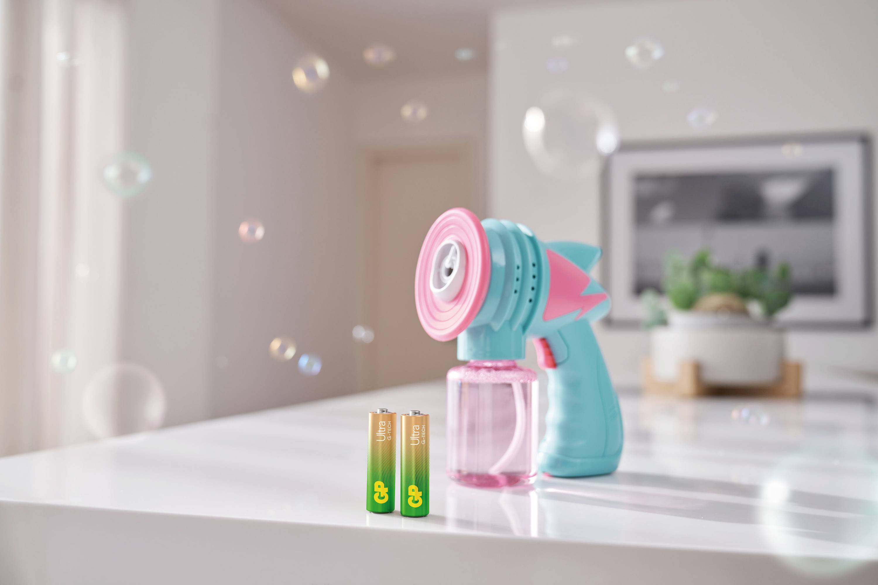 Machine à bulles de savon bleue et rose posée sur une table. Un tableau est accroché au mur en arrière-plan et une plante est présente. Deux piles sont disposées à côté.