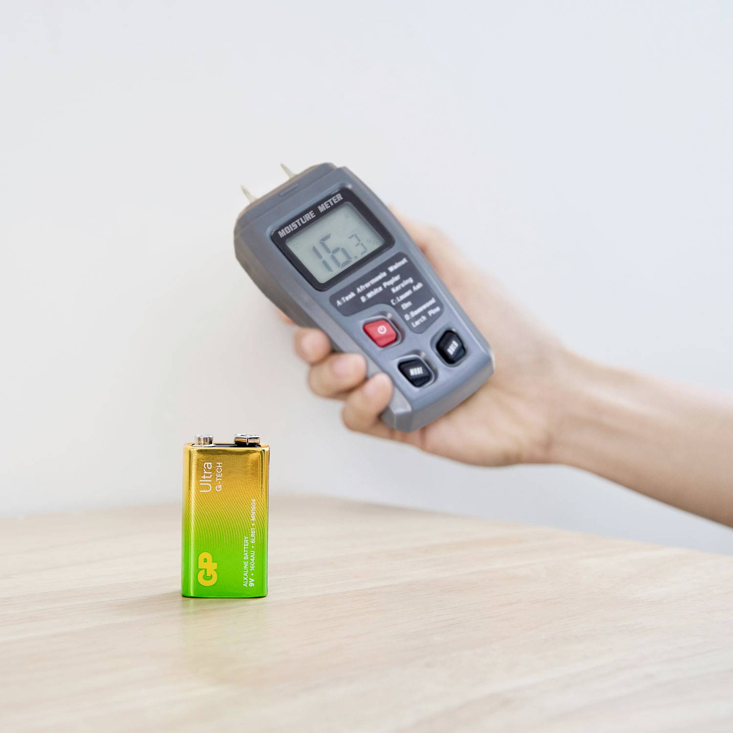La main tient un humidimètre avec un affichage de 16,3 dirigé vers une batterie de 9 volts; mesure probablement l'état de la batterie.