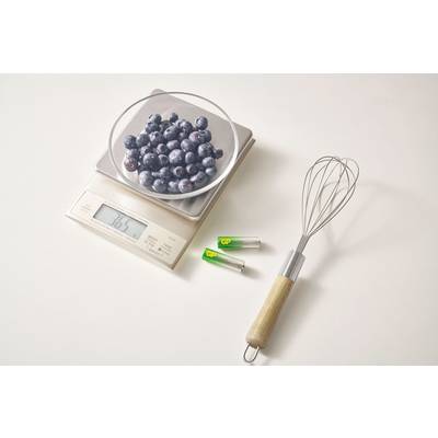 Une balance de cuisine avec un bol rempli de myrtilles, à côté un fouet et deux piles AA sur un fond clair.