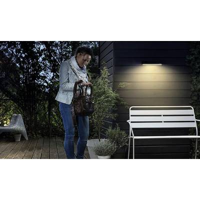 Une personne se tient la nuit devant un banc éclairé sur une terrasse et regarde dans un sac à main. Des plantes sont en arrière-plan.