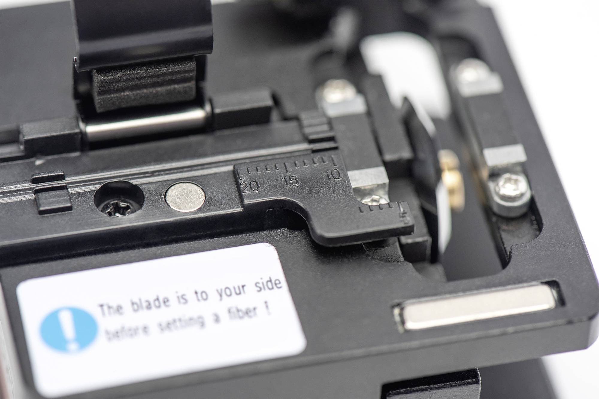 Close-up of a fiber optic cutter with scale and instruction label 'The blade is to your side before setting a fiber!'