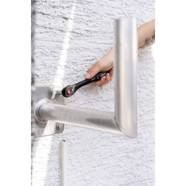 Une personne resserre un boulon sur un tuyau métallique fixé à un mur blanc texturé à l'aide d'une clé à molette noire.