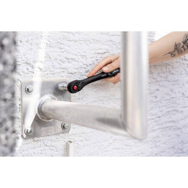 Une personne utilise une clé à molette pour resserrer un boulon sur une rambarde métallique fixée à un mur texturé. Un tatouage est visible sur l'avant-bras de la personne.