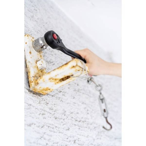 Une main serre une clé à cliquet pour resserrer ou desserrer un boulon rouillé sur une pièce métallique fixée à un mur avec des câbles attachés.