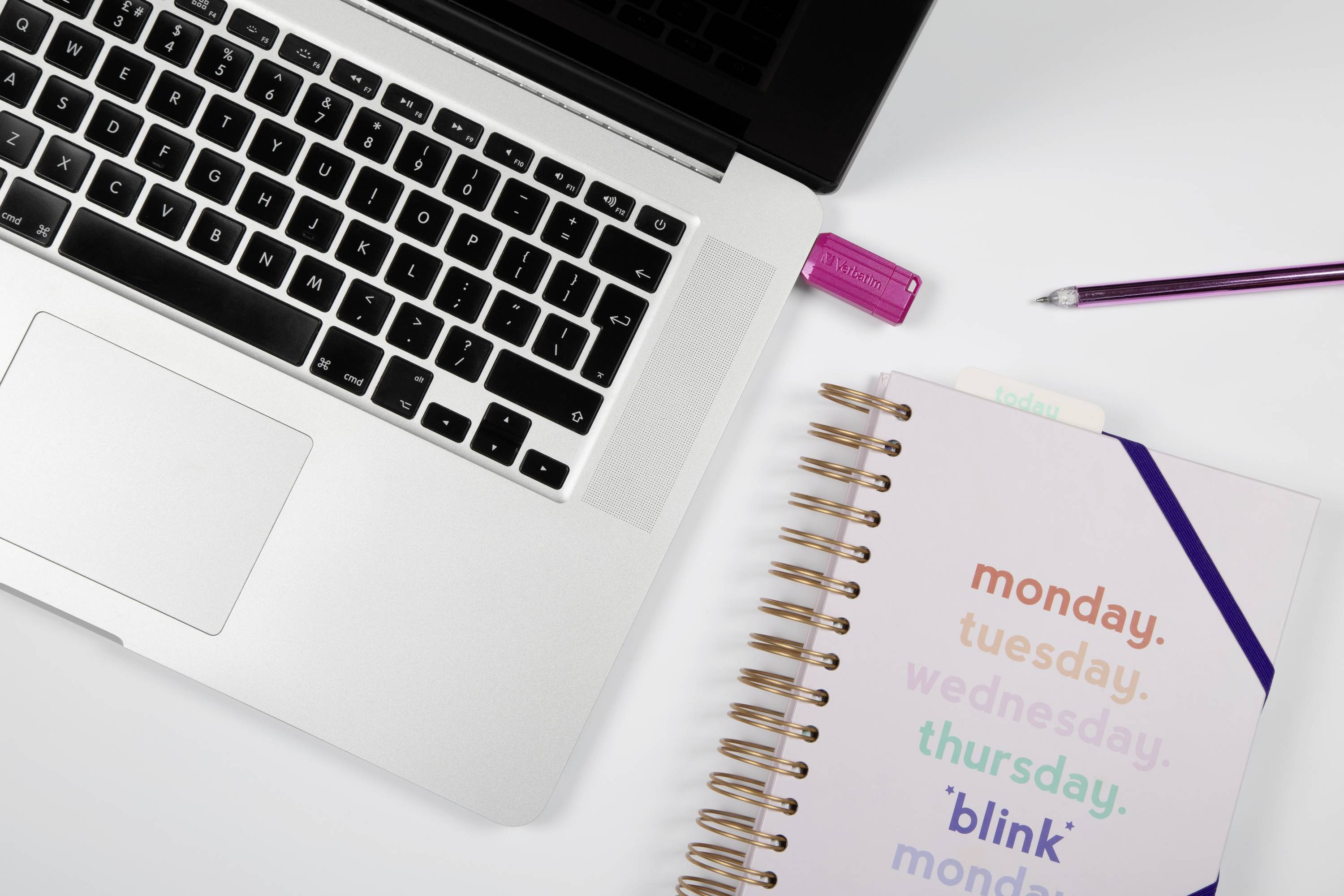 Un ordinateur portable sur une table. À côté, un carnet avec les jours de la semaine et une clé USB. Un stylo violet dans le coin supérieur droit.