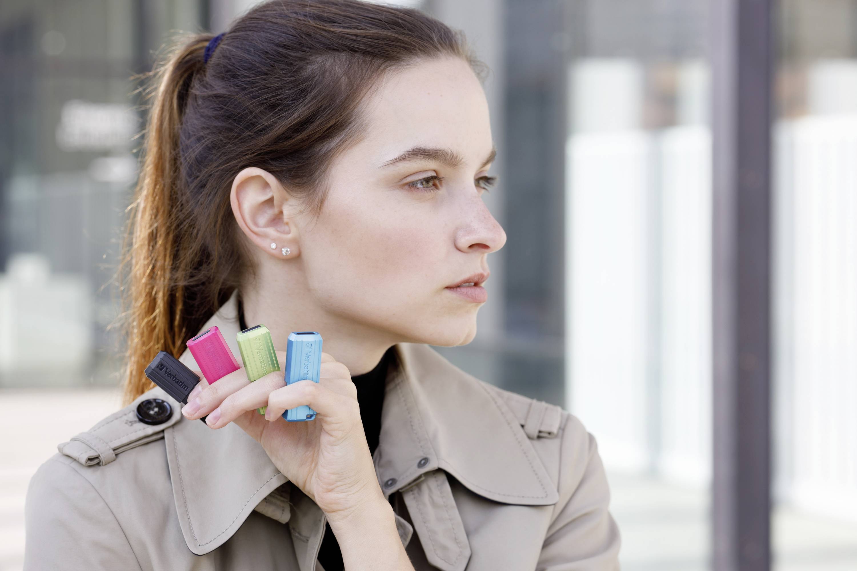 Une femme de profil tient quatre objets rectangulaires colorés. Elle porte un trench-coat et paraît pensive.