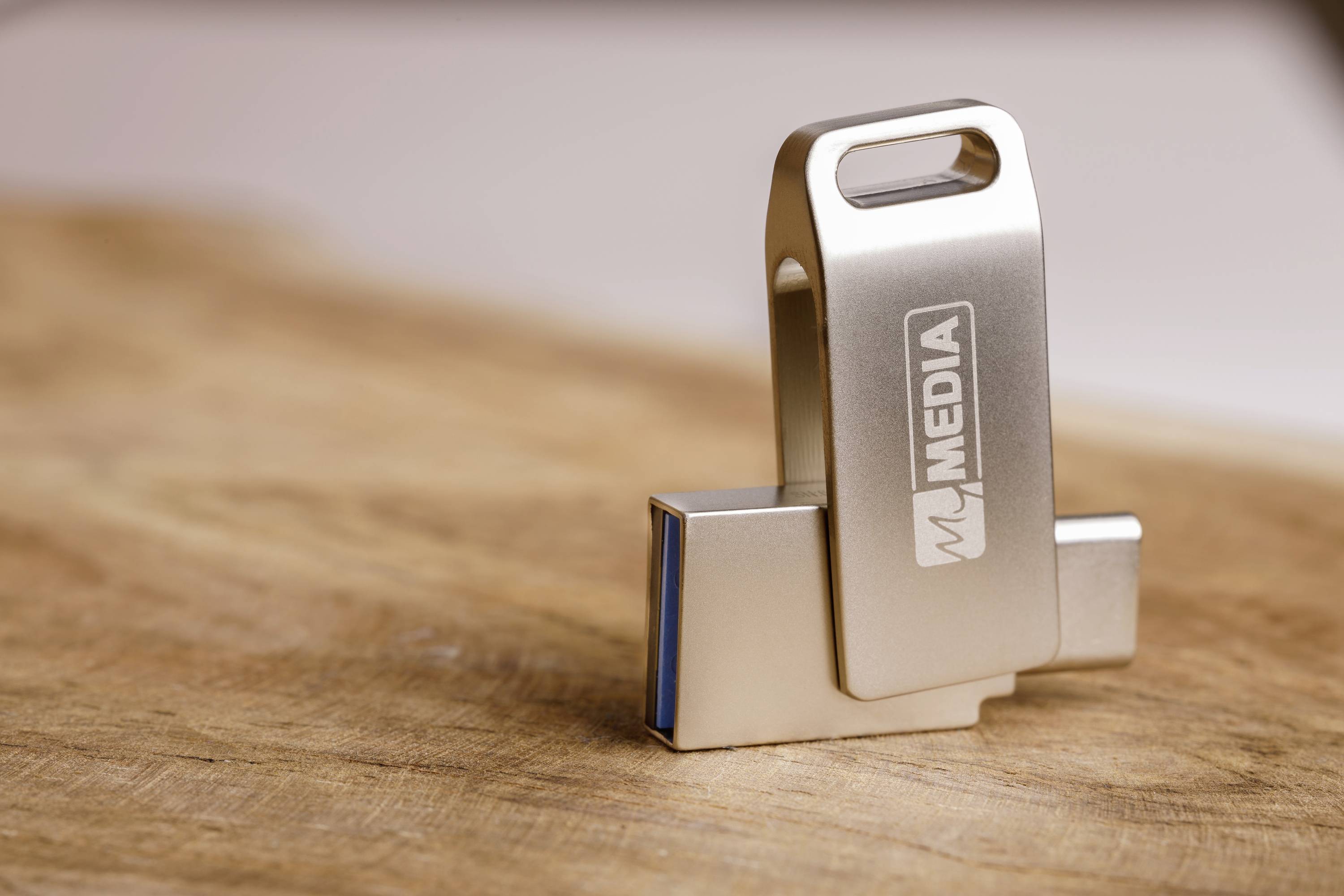 Une clé USB avec le logo 'MJ MEDIA' est posée sur une surface en bois. La clé, en métal, est partiellement dépliée.