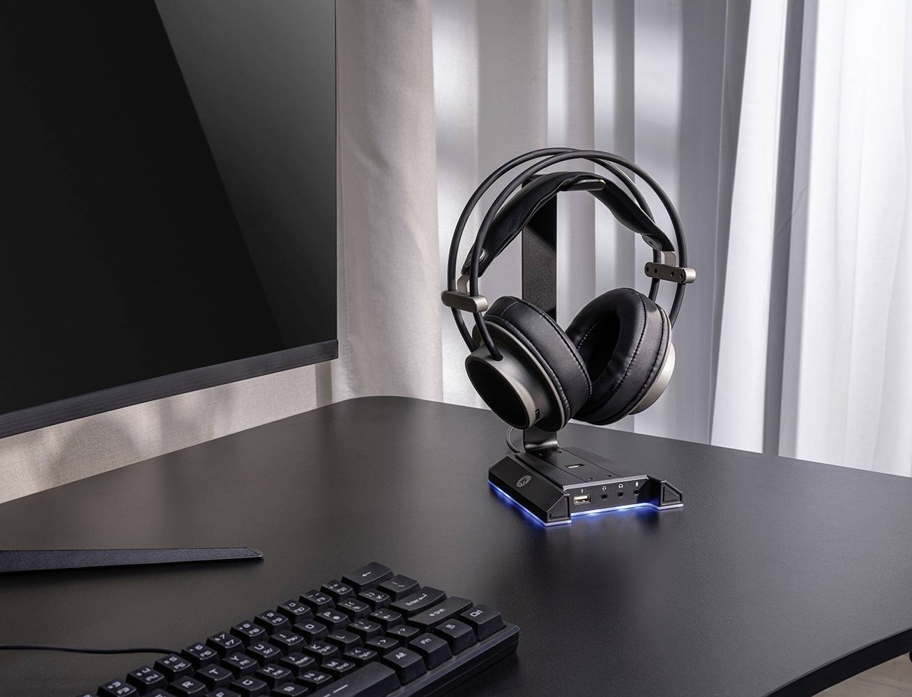 Un bureau avec un écran, un clavier et une paire d'écouteurs sur un support. La lumière du soleil pénètre à travers le rideau.