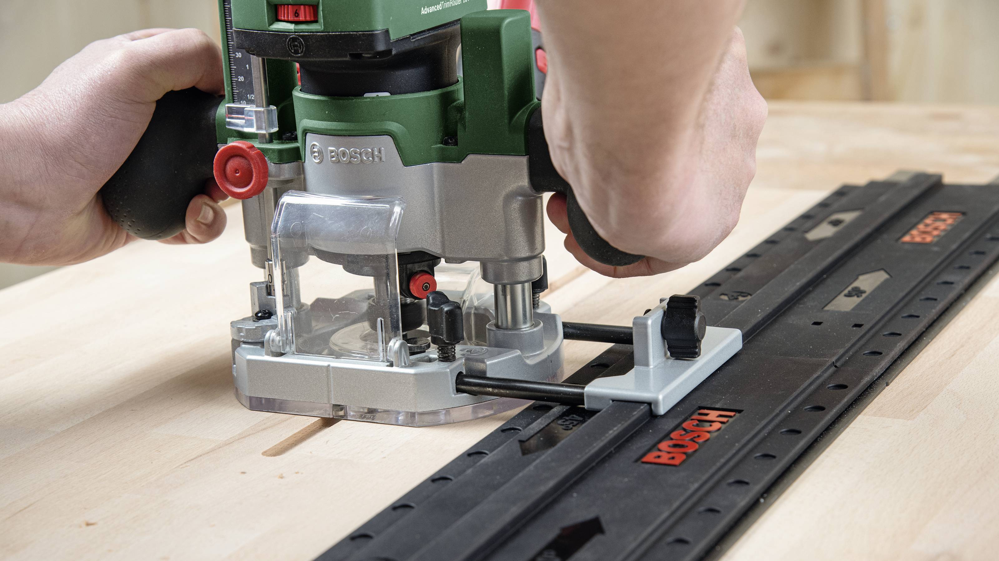 Une défonceuse Bosch est utilisée sur un rail de guidage pour fraiser une pièce de bois. Les mains tiennent la défonceuse pour un travail précis.