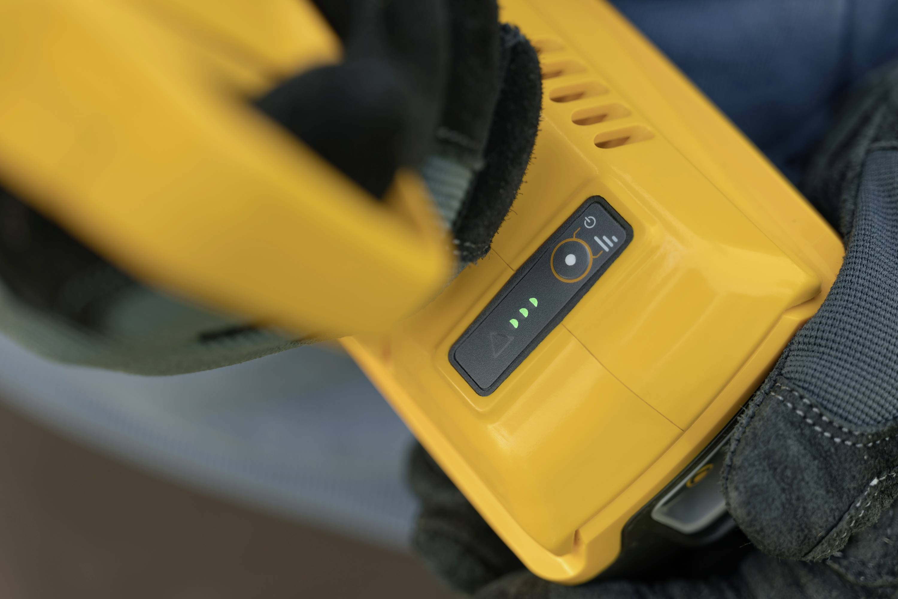 Batterie d'outil jaune en gros plan avec des voyants LED verts indiquant le niveau de charge. Une personne tient la batterie avec des gants de travail.