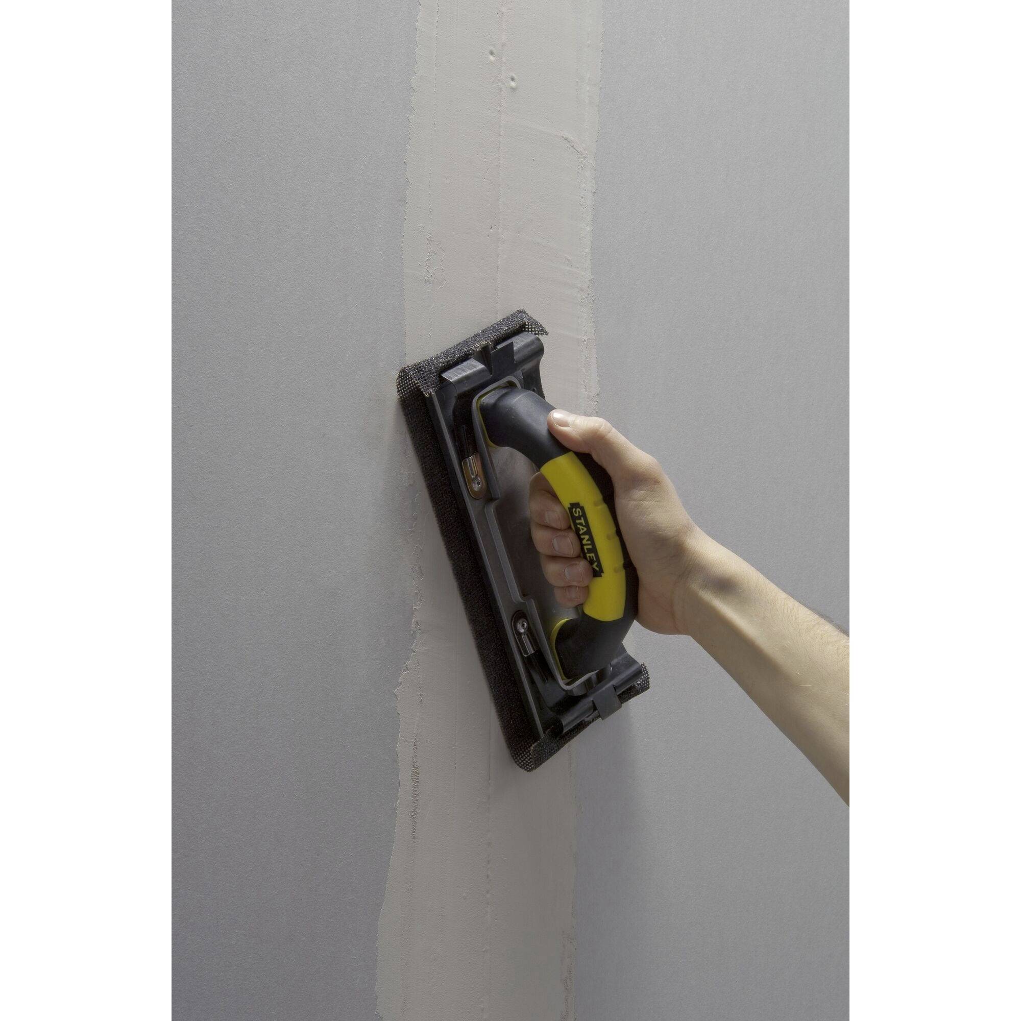 Poncage d'un mur en plaque de plâtre à la main avec un bloc à poncer et une poignée jaune.