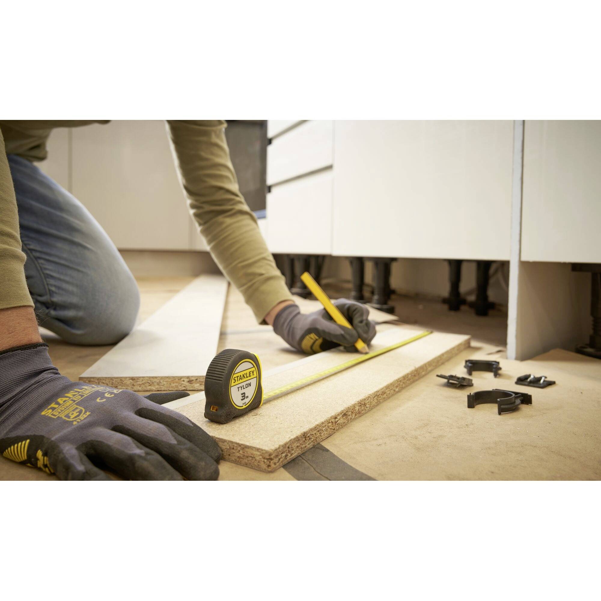 Une personne mesure et marque un morceau de bois avec un mètre ruban. Sur le sol sont disposés des vis et des outils utilisés pour le travail.
