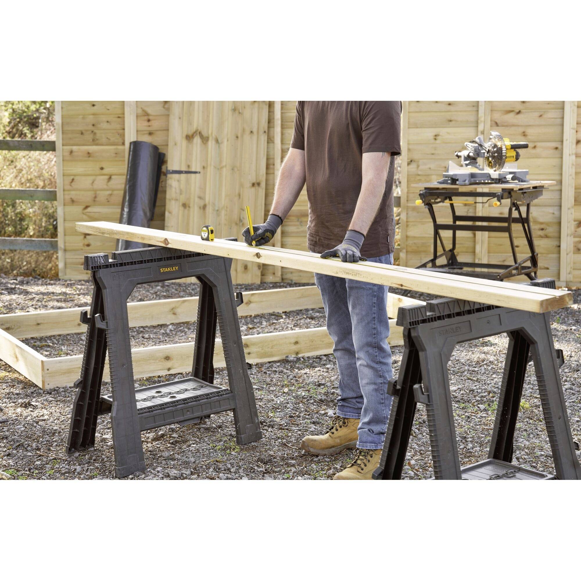 Une personne mesure une planche de bois avec un mètre ruban dans un atelier extérieur. Une scie est visible sur une table en arrière-plan.
