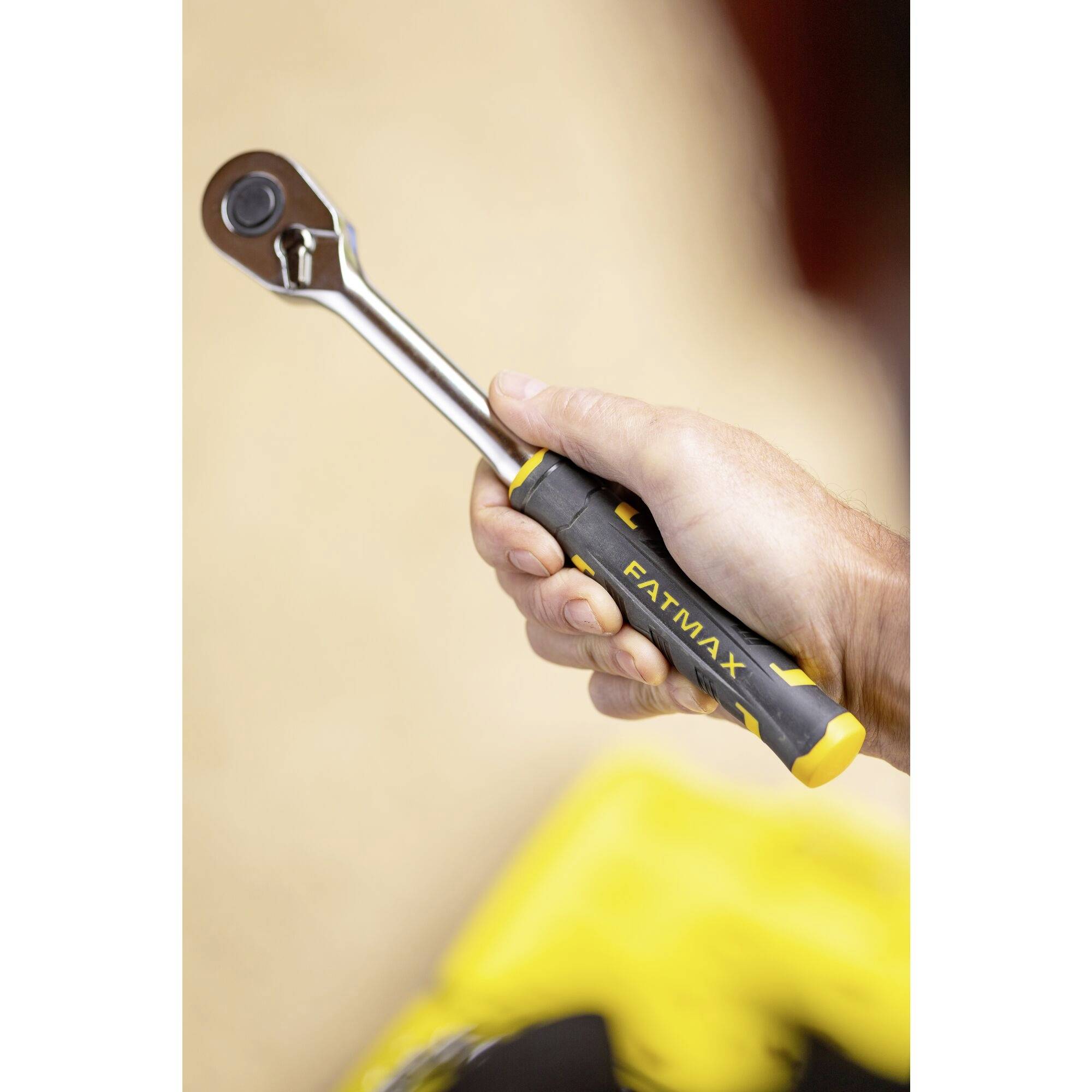 Une main tient une clé à cliquet avec une poignée noire et jaune sur laquelle est inscrit 'FatMax'. À l'arrière-plan, des outils jaunes sont visibles de manière floue.