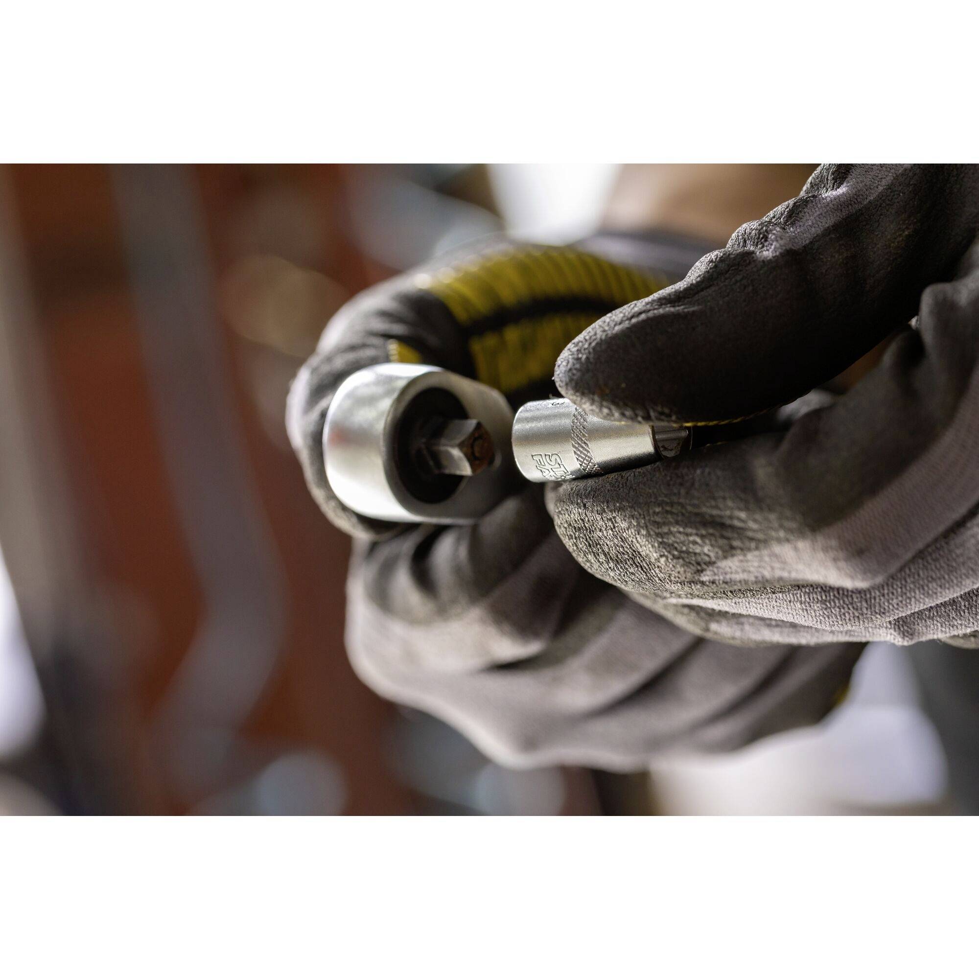 Une main gantée de gris tient un outil métallique à vis ; des éléments industriels flous sont visibles en arrière-plan.