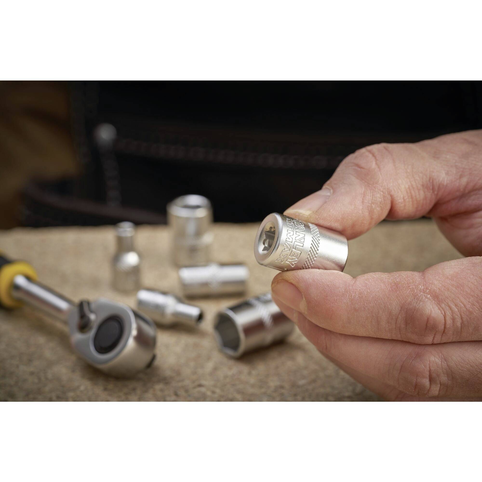 Une main tient un écrou à souder, entouré d'autres outils, sur une table. Une boîte à cliquet est visible en arrière-plan.