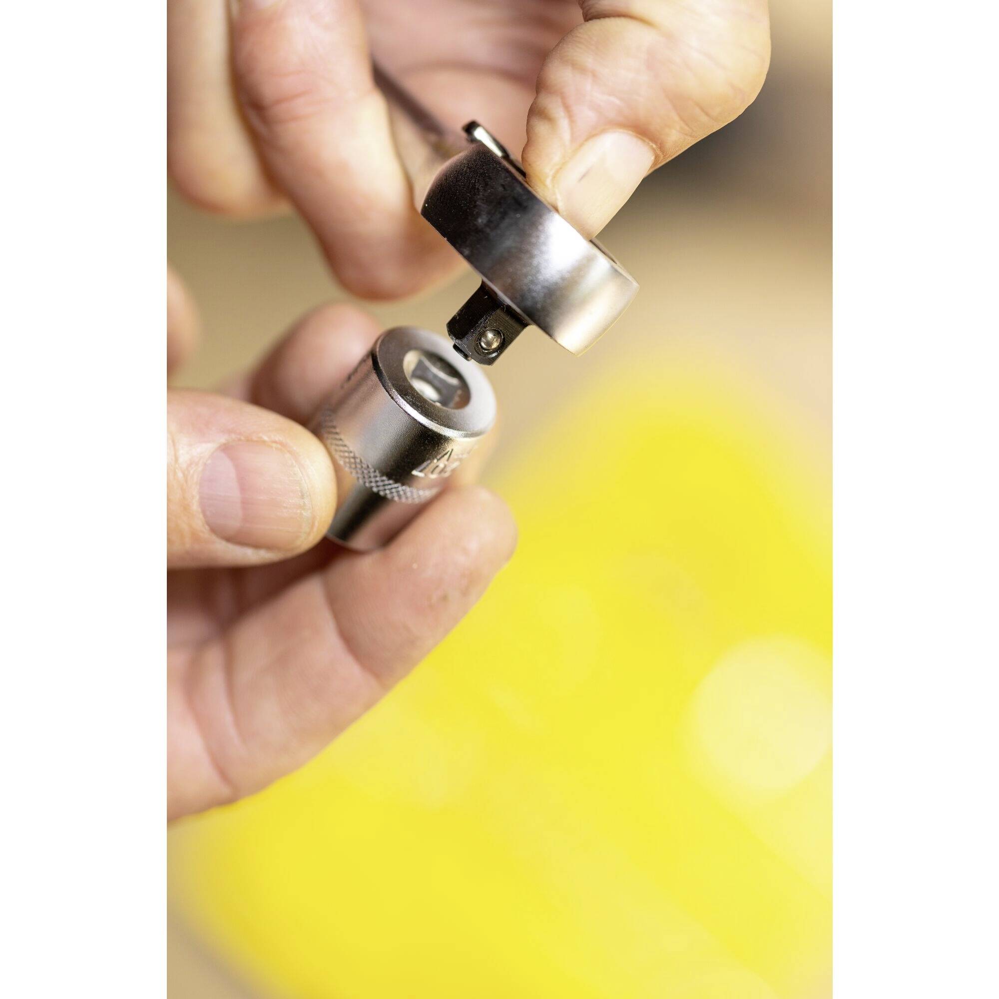 Gros plan sur deux mains manipulant un composant métallique, probablement un élément de connexion métallique. L'arrière-plan est flou et de couleur jaune.