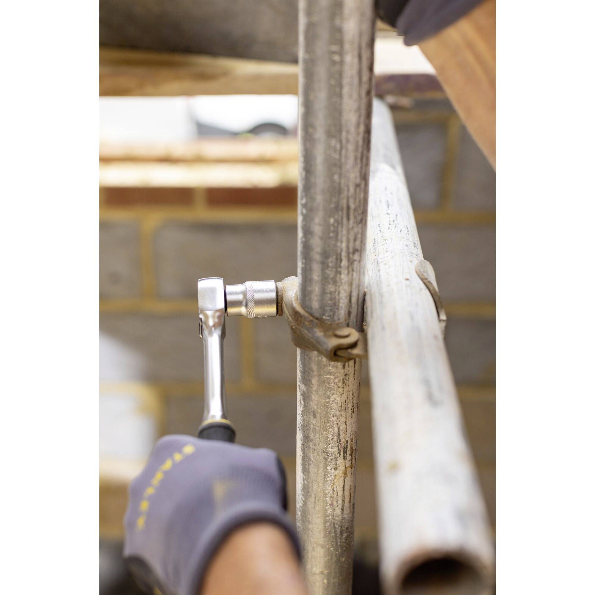 Une personne portant des gants utilise une clé à molette pour serrer un tube d'échafaudage. Un mur de briques est visible en arrière-plan.