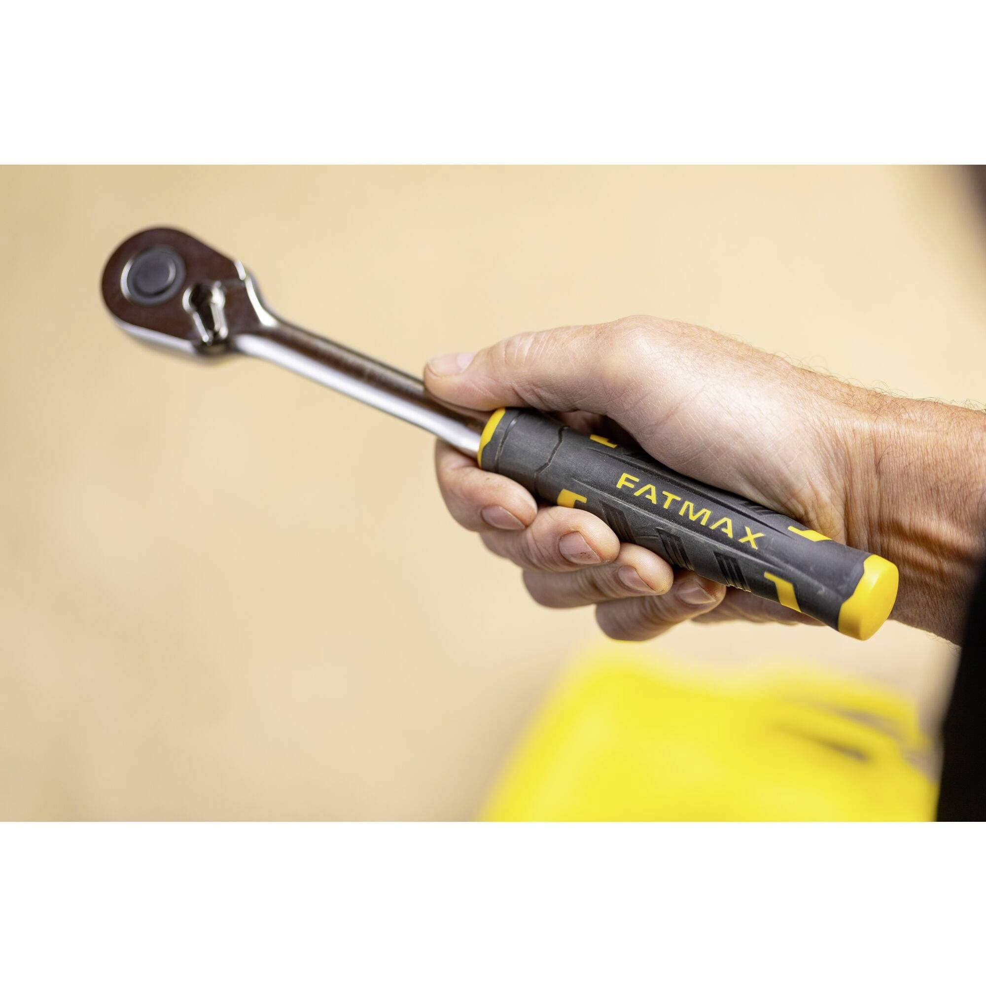 Une main tient une clé à cliquet portant l'inscription 'FATMAX', avec un sac jaune flou en arrière-plan.