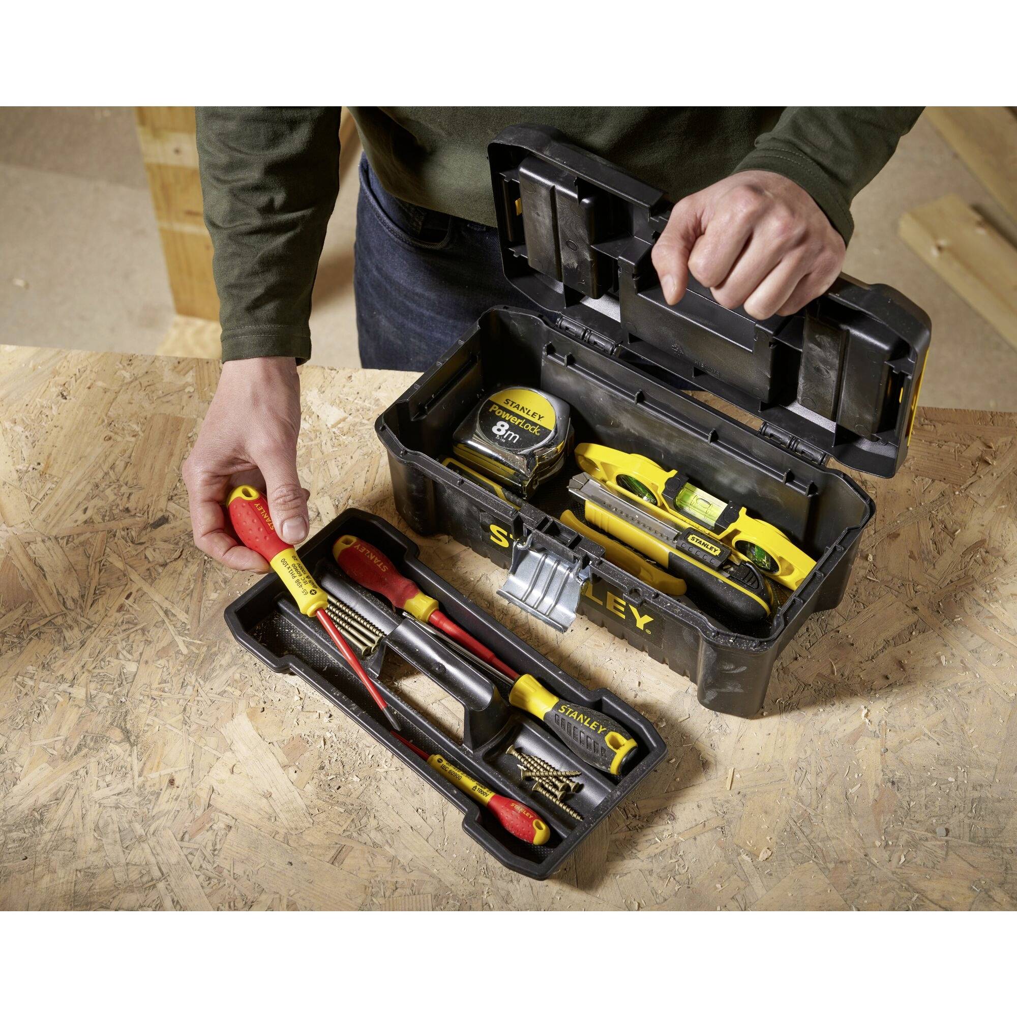 Une personne ouvre une boîte à outils posée sur une table, contenant divers outils tels que des tournevis, un mètre ruban et un niveau à bulle.