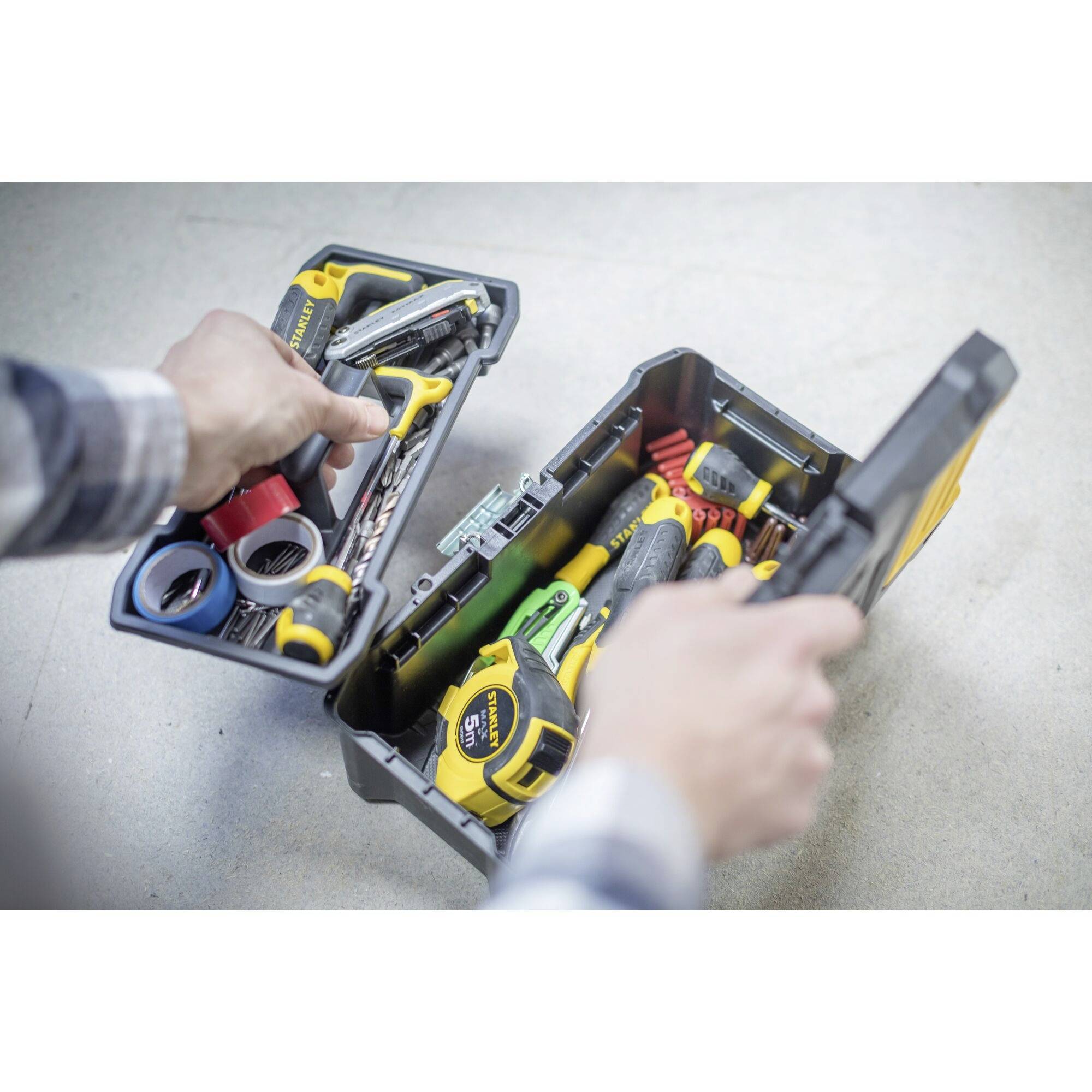 Une personne ouvre une boîte à outils remplie d'outils, comprenant un mètre ruban, des tournevis et des pinces sur un fond gris.