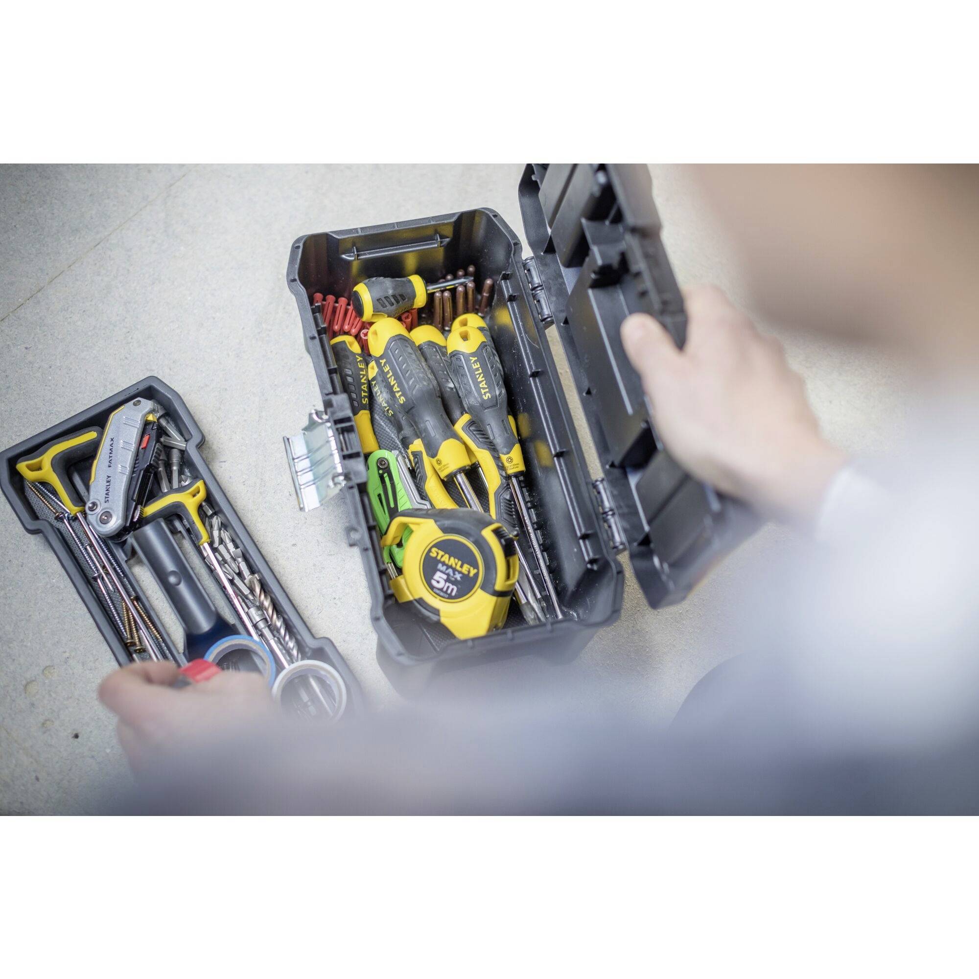 Une boîte à outils ouverte contenant divers outils tels que des tournevis, un mètre ruban et des vis est observée par une personne.