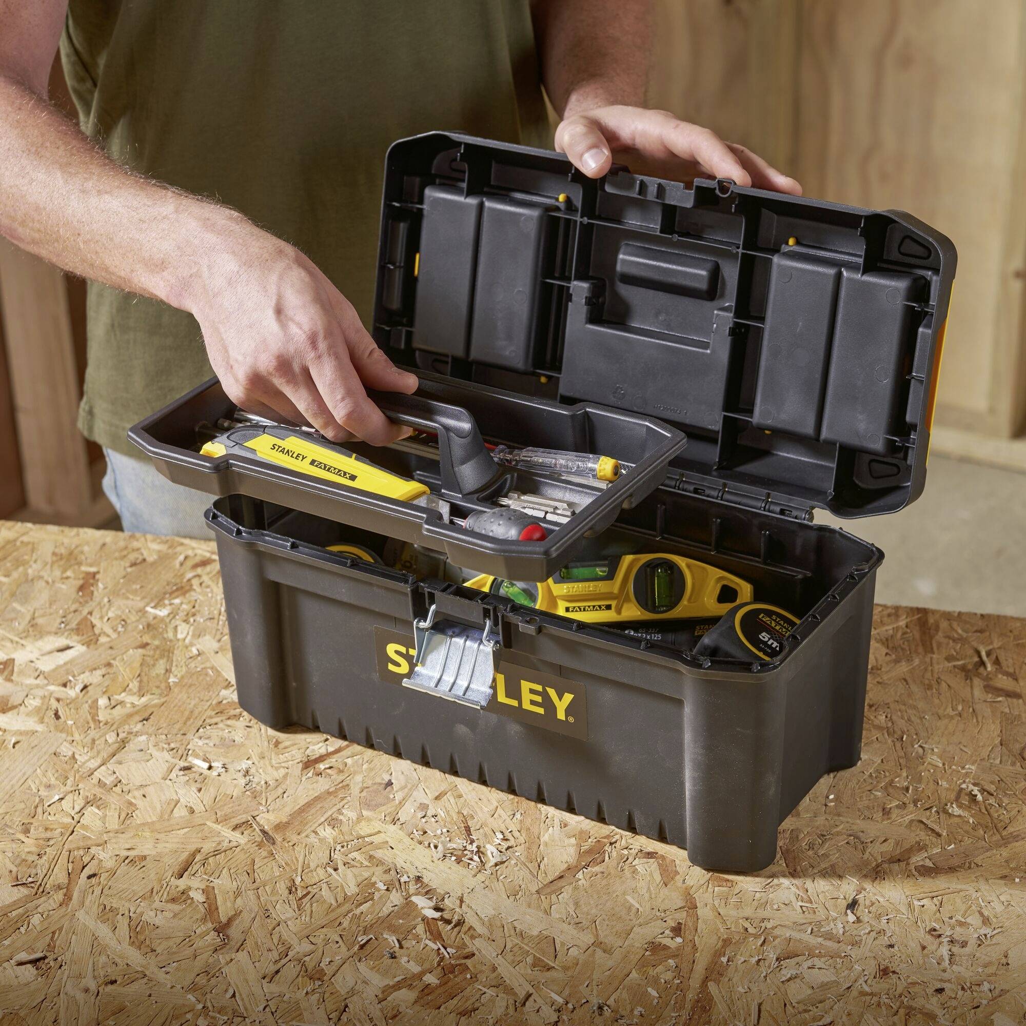 Un homme ouvre une boîte à outils remplie de divers outils et d'un mètre ruban, posée sur une table en bois.