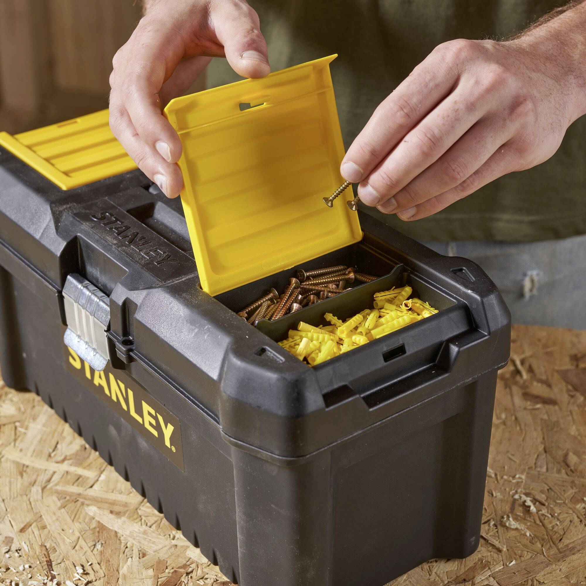 Une personne ouvre le couvercle d'une boîte à outils noire avec un tiroir jaune pour retirer des vis et des chevilles sur une surface en bois.