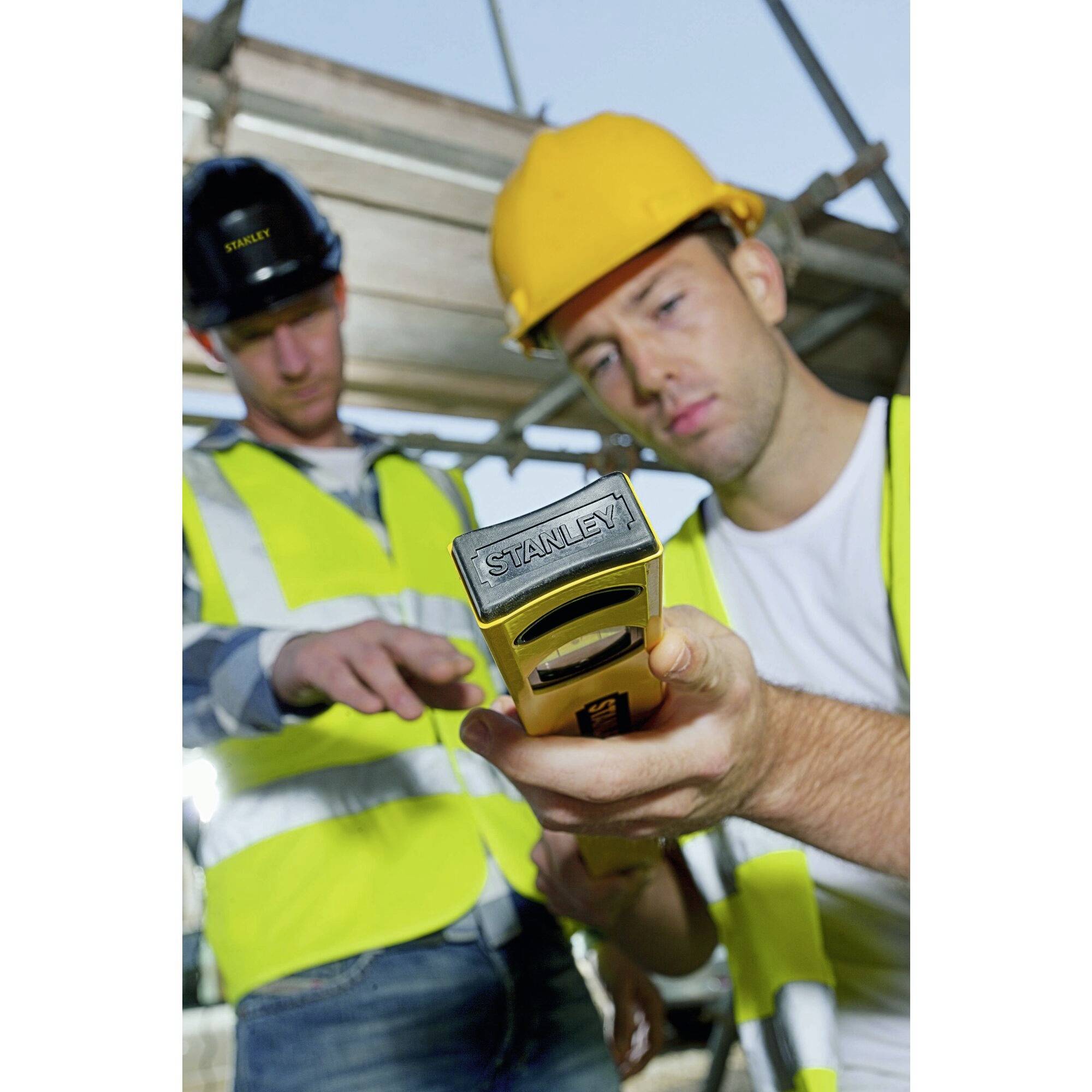 Deux ouvriers du bâtiment en gilets de sécurité et casques travaillent sur un projet de construction. L'un tient un outil à la main, tandis que l'autre pointe du doigt.