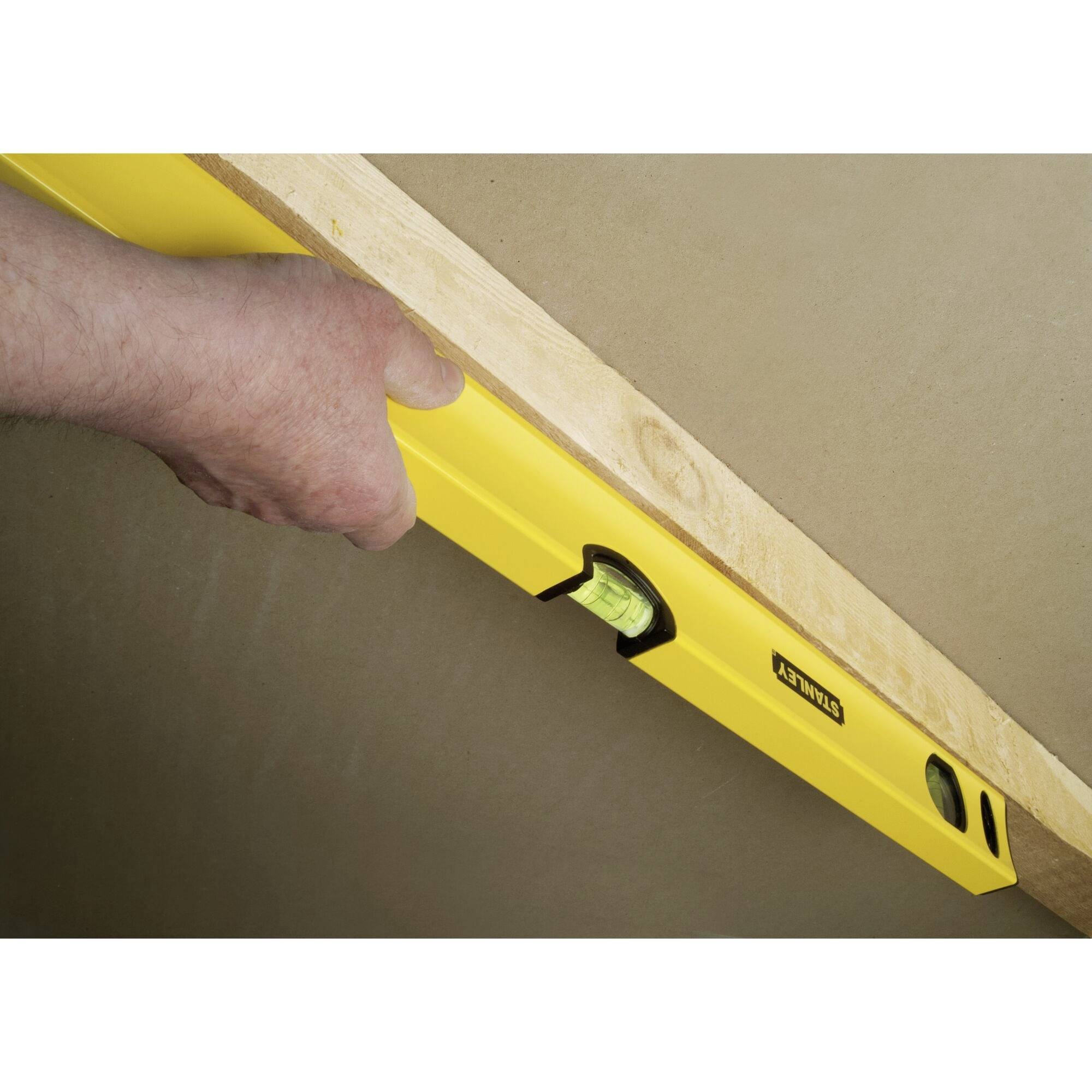 Une main tient un niveau à bulle jaune contre une planche en bois pour vérifier l'alignement. Le niveau indique si la planche est droite.