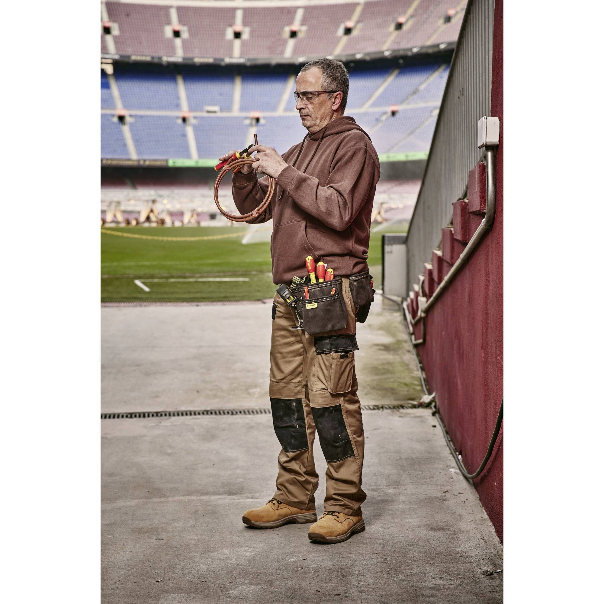 Un ouvrier se tient dans le stade et travaille sur un câble. Il porte des vêtements de travail et des outils à sa ceinture.
